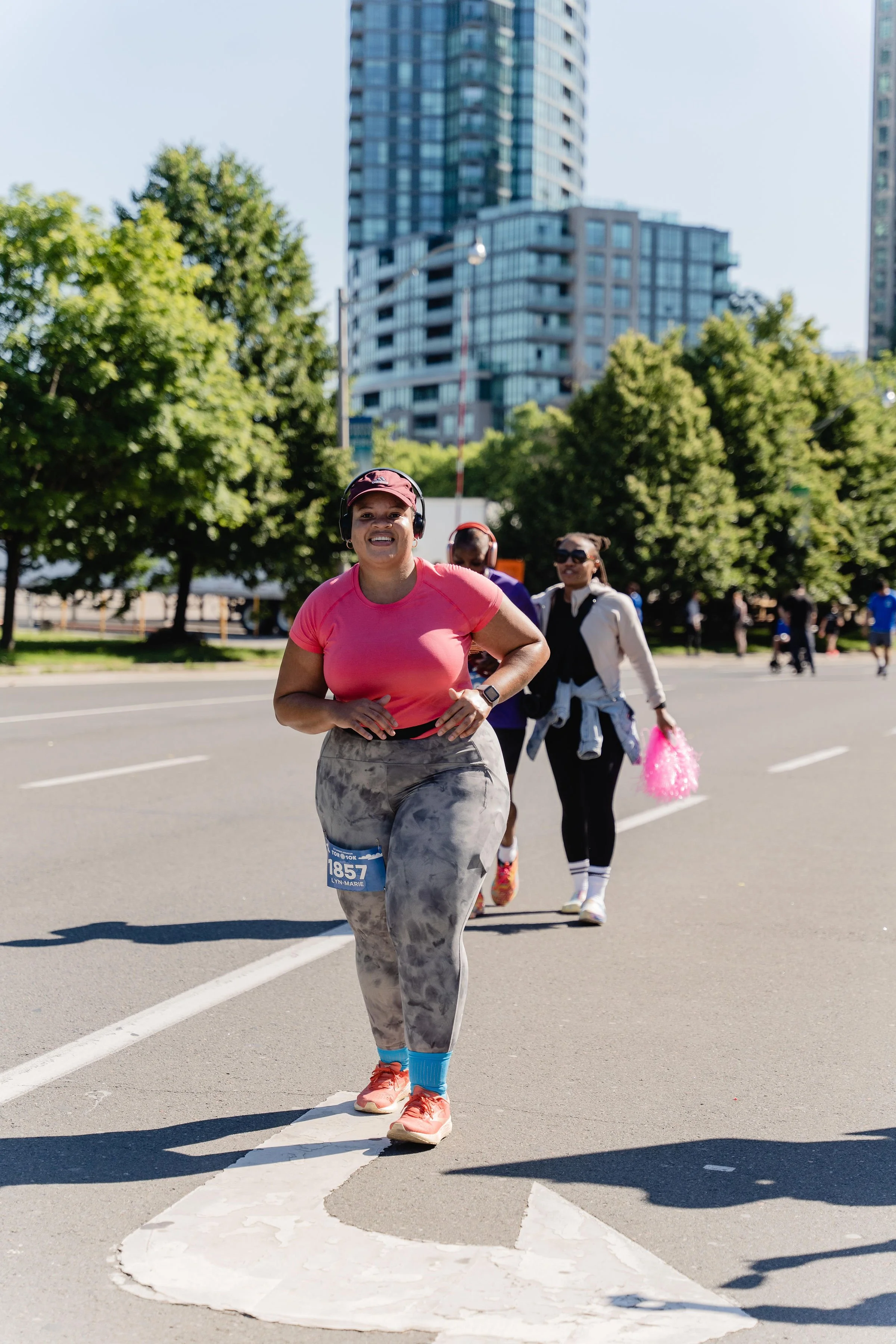 Female runner smiling and running in a road race, wearing a pink shirt, gray tie-dye leggings, and a red cap, with others running behind her in an urban setting with trees and tall buildings.