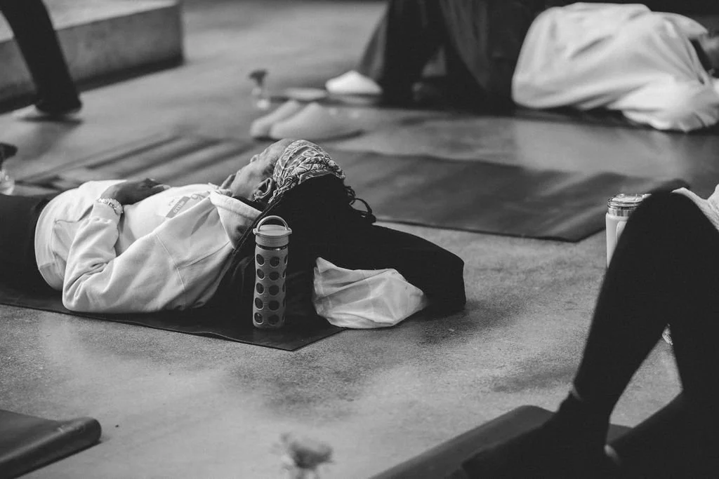A person lying on their back on a yoga mat in a meditation or relaxation pose at a yoga studio or gym, with a water bottle beside them.