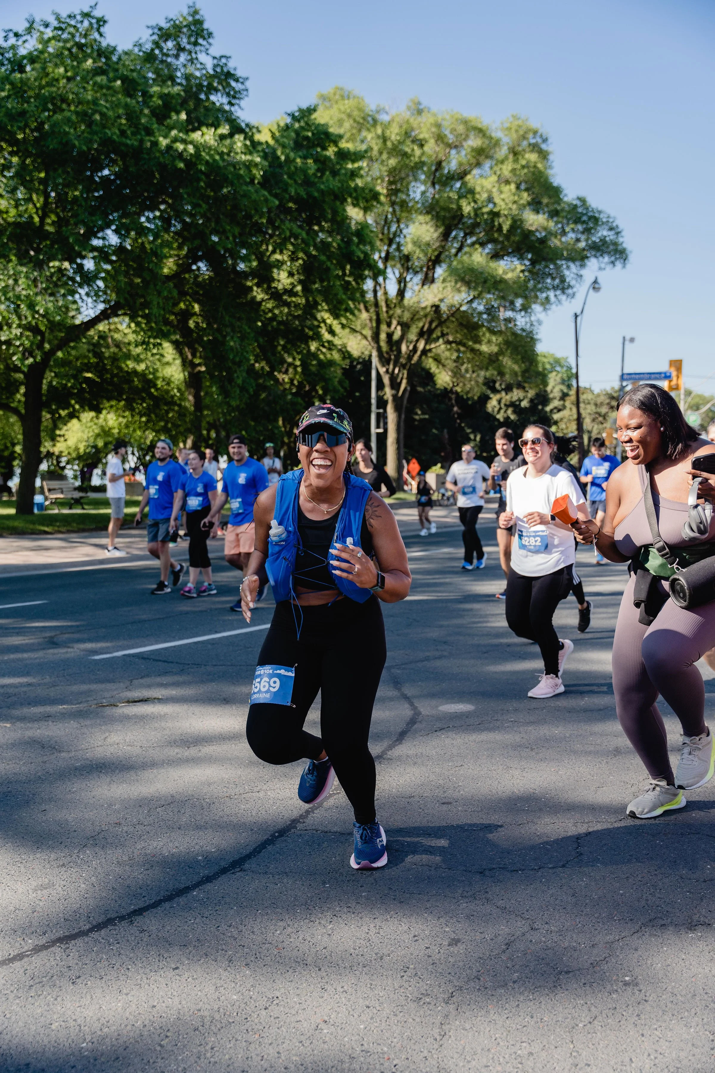 Women participating in a road race, running and smiling under a clear blue sky with trees in the background.
