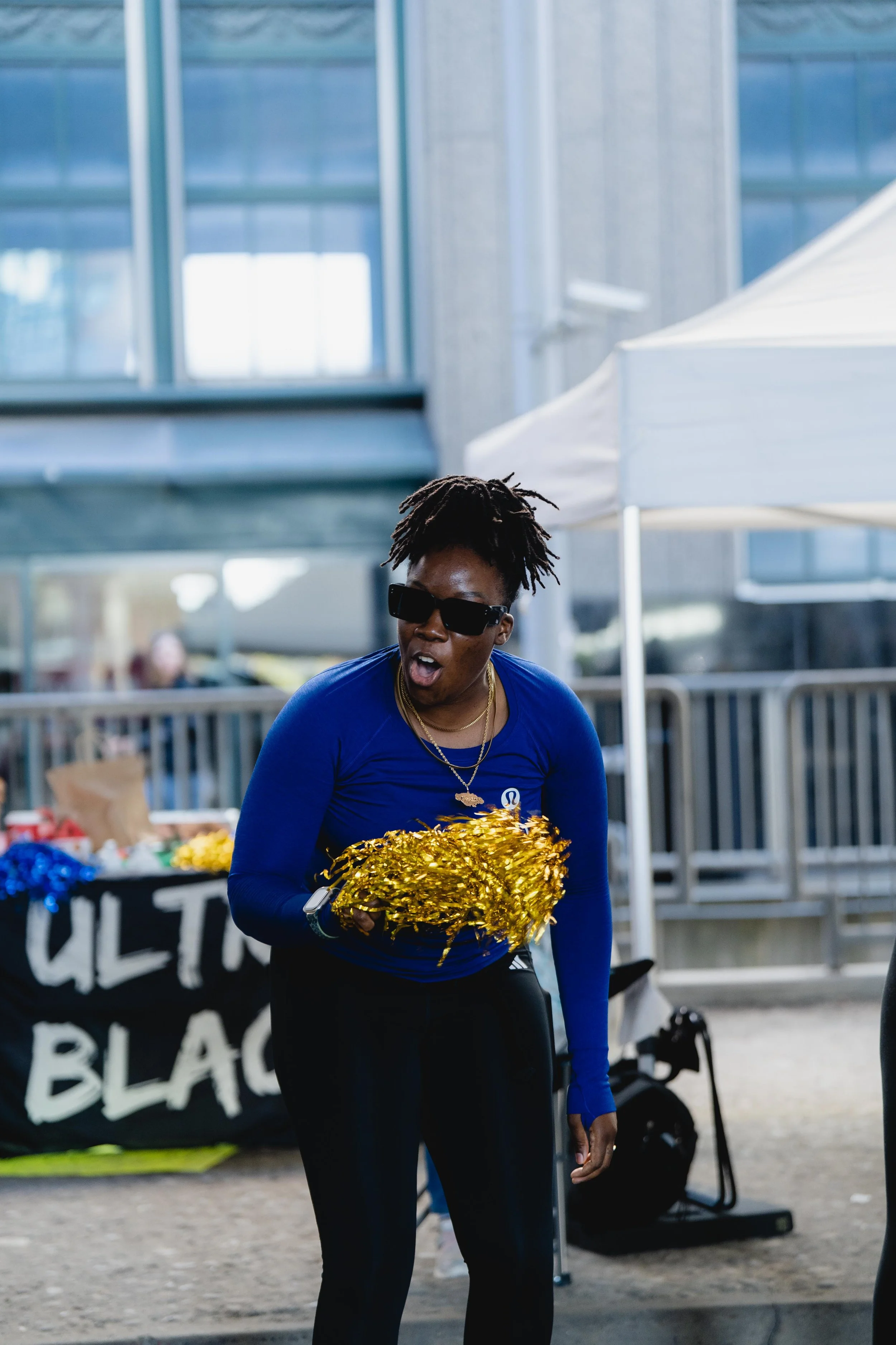 A woman wearing sunglasses and blue athletic clothing holding gold pom-poms at an outdoor event.