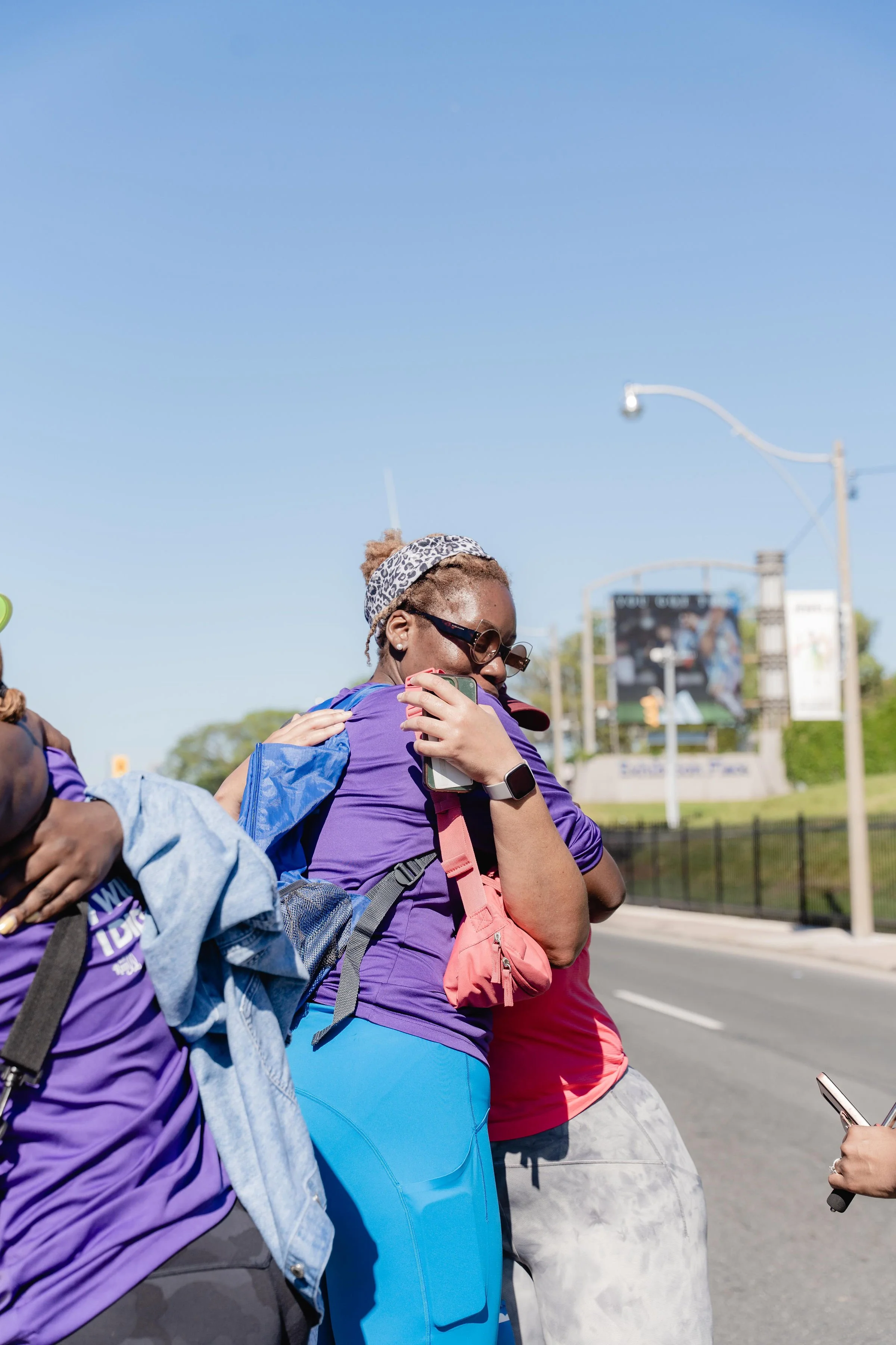 Two women hugging outdoors on a sunny day near a road with trees in the background. One is wearing sunglasses and a leopard-print headband, and the other is holding a phone.