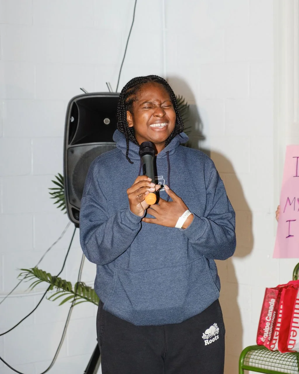 A young woman with braided hair wearing a blue hoodie singing passionately into a microphone, with a speaker and some plants in the background.