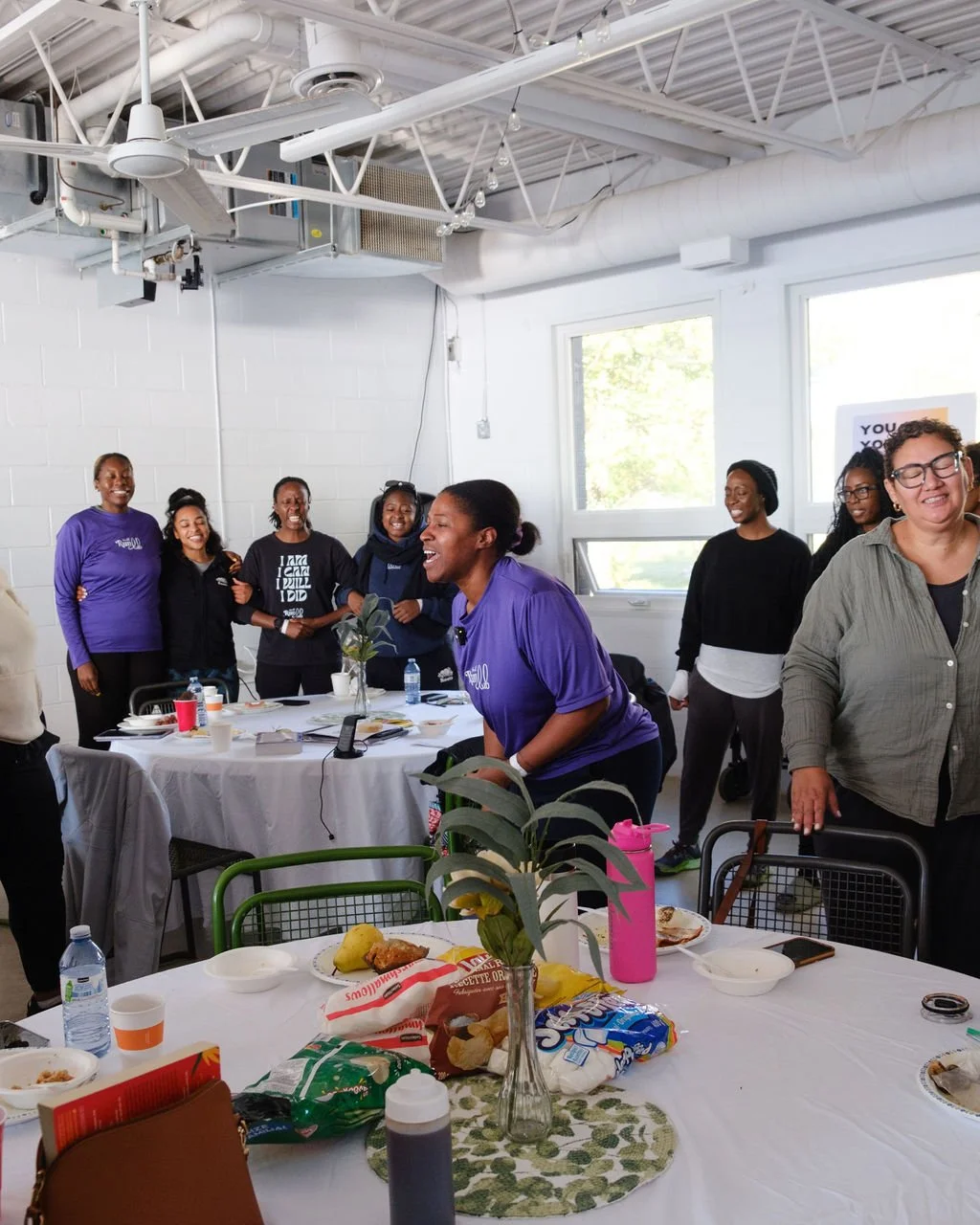 A group of women gathered in a bright room, smiling and laughing. Some are standing, some are sitting at tables with food, drinks, and a flower centerpiece. The room has large windows and industrial ceiling features.