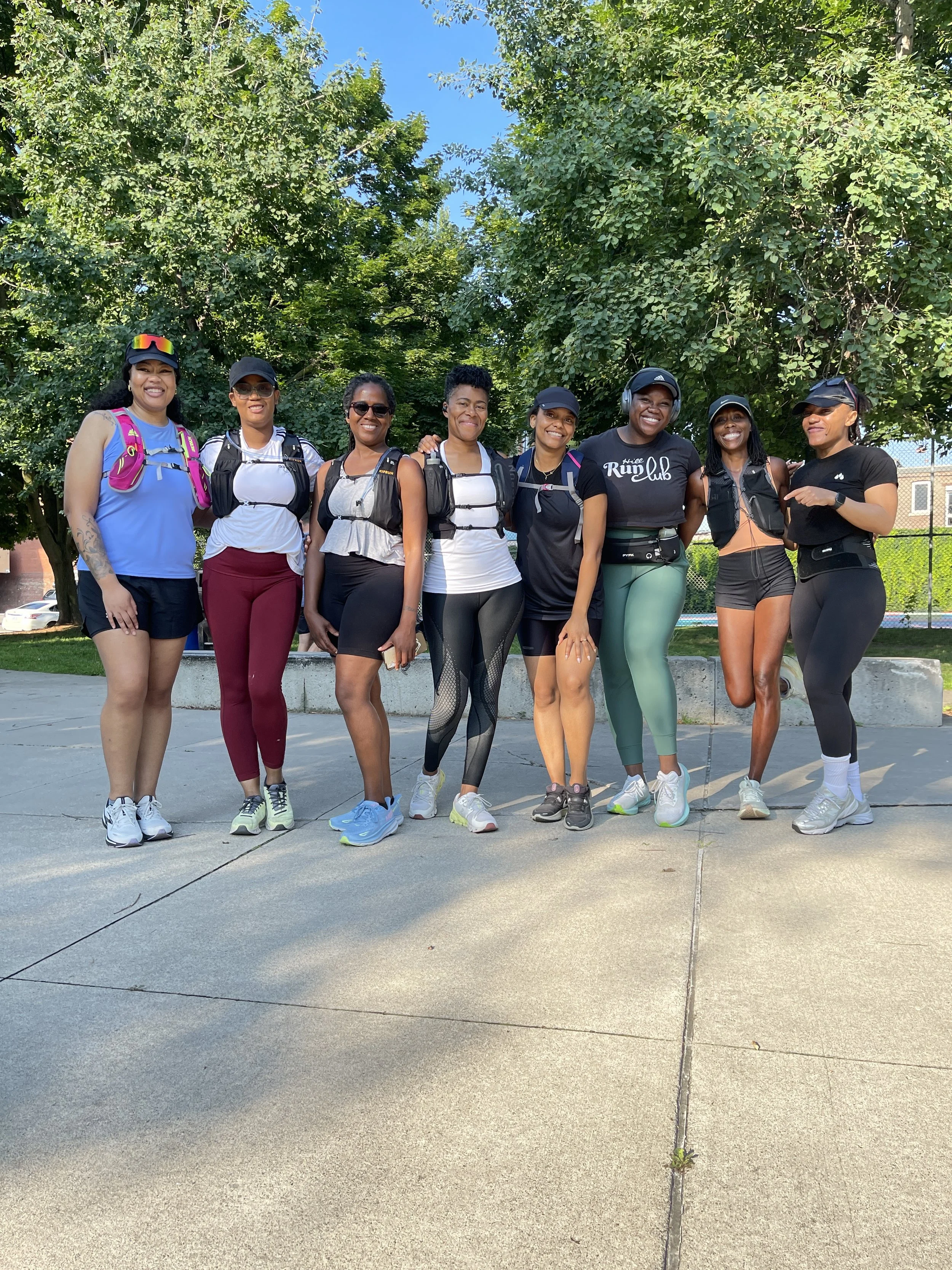 Group of nine women standing together outdoors on a sunny day, dressed in athletic wear and sneakers, smiling at the camera with trees and a clear blue sky in the background.
