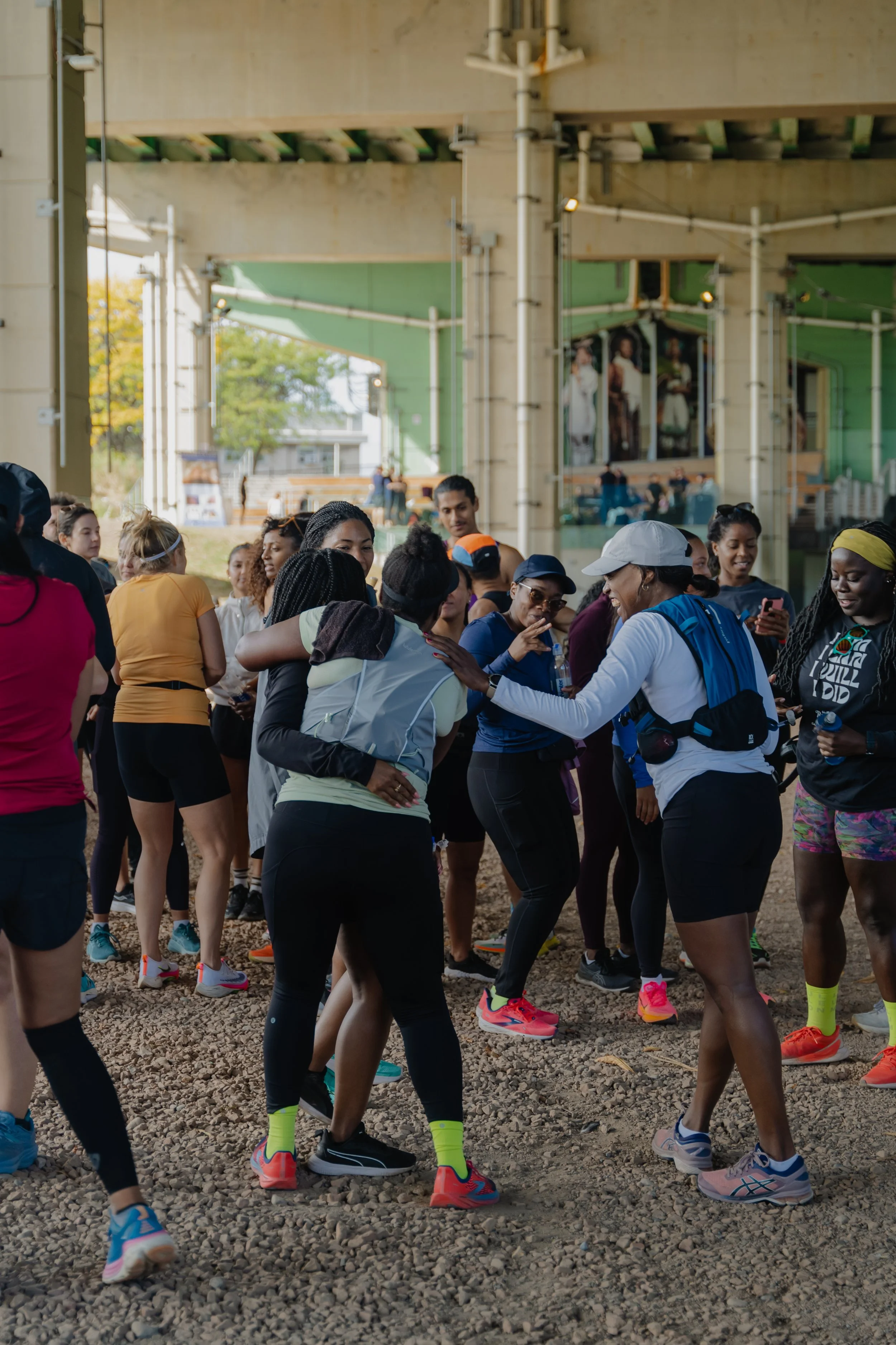 Women and men dressed in athletic wear are gathered underneath an overpass, engaging in friendly interactions and preparations for a run or outdoor exercise event.