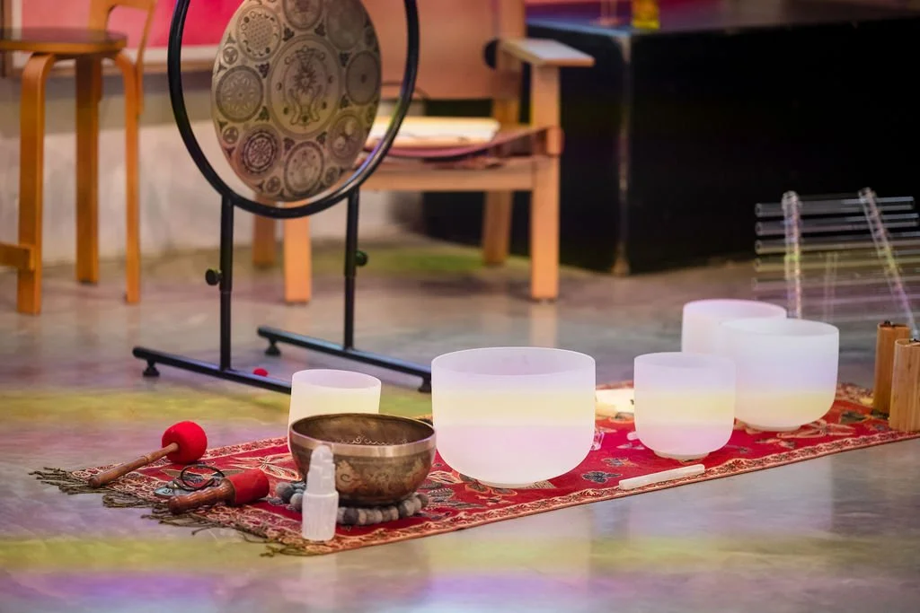 A collection of singing bowls, a mallet, and a spray bottle on a red patterned rug in a room with chairs and a gong in the background.