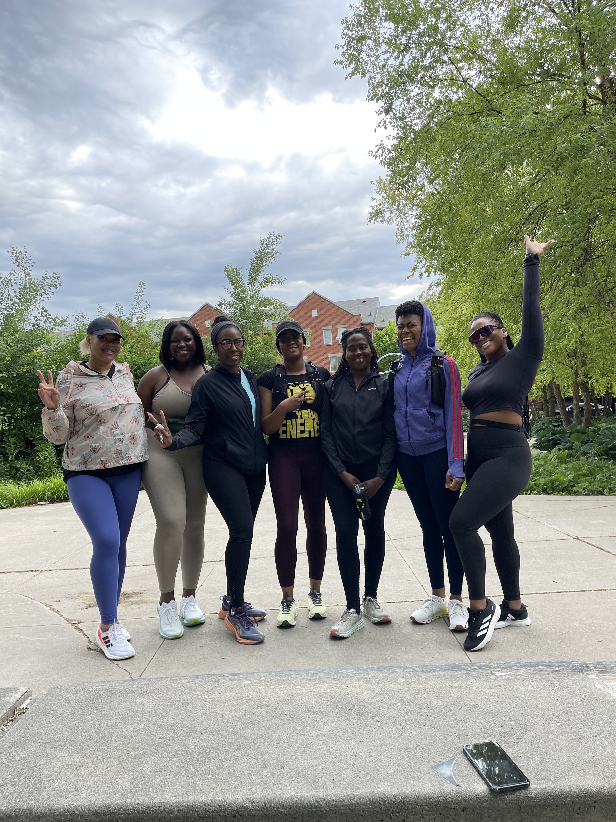 A group of seven women standing outdoors on a sidewalk, smiling and posing for a photo, with trees and residential buildings in the background on a cloudy day.