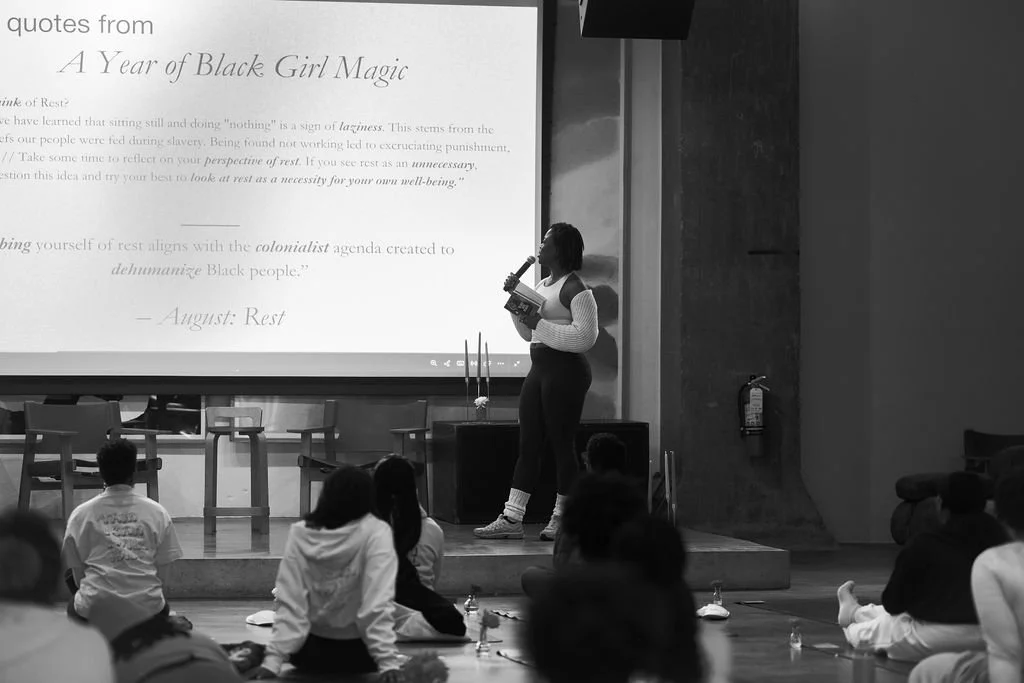 Woman in a white and dark outfit, holding a book, standing on a stage in front of a large screen with text, addressing an audience seated on the floor.