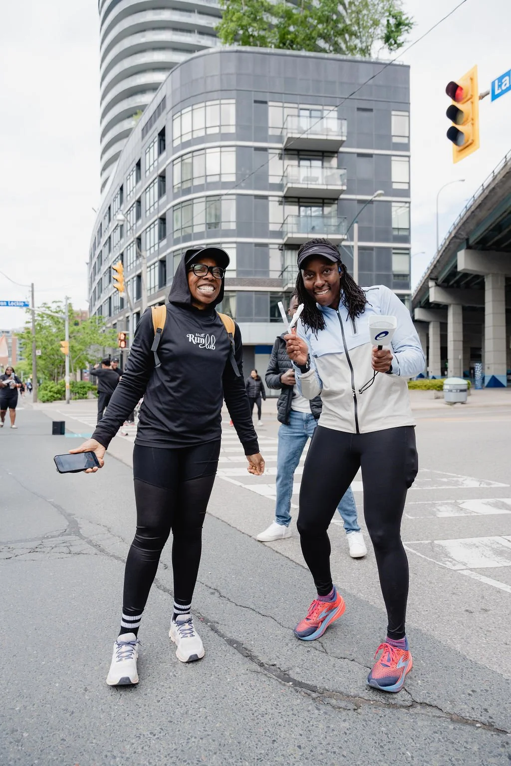 Two women standing at a crosswalk in an urban area, smiling at the camera. They are dressed in athletic clothing, with buildings, traffic lights, and a few other people in the background.