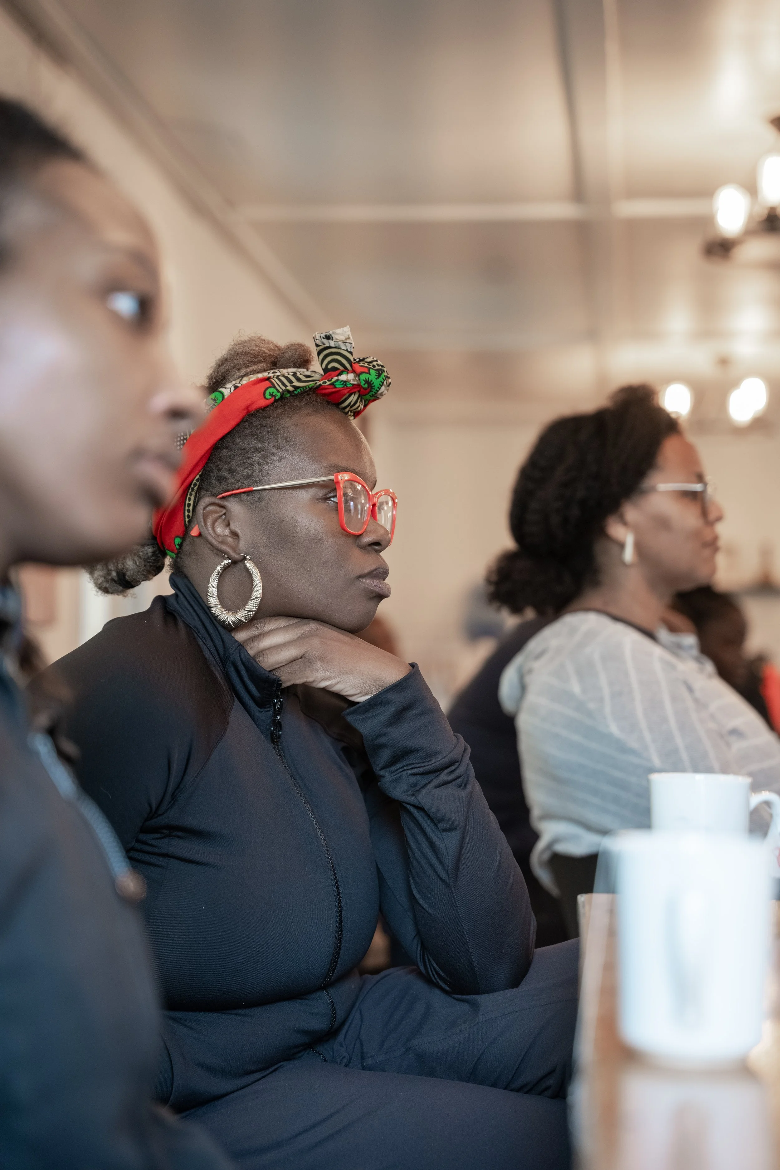 A woman with large hoop earrings, red glasses, and a colorful headwrap attentively listening at a meeting or conference, with other women seated beside her.
