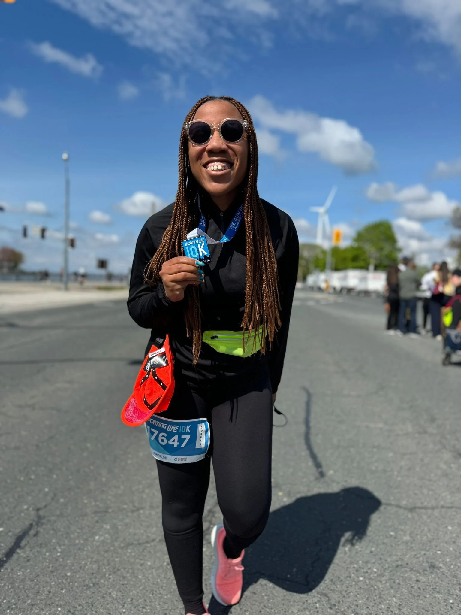 Woman smiling, running in a race, holding a medal, wearing black athletic clothing, pink running shoes, race bib, sunglasses, with wind turbines under a blue sky.