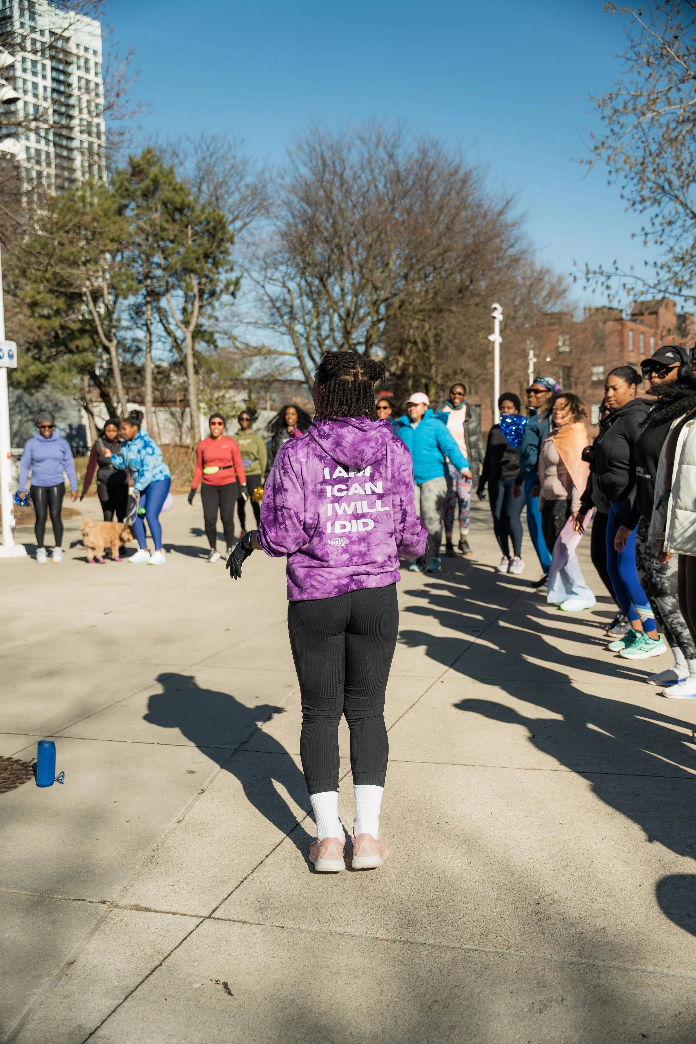 A group of people gathered outdoors in a park or plaza, with a woman in the foreground wearing a purple tie-dye hoodie that says 'I AM, I CAN, I WILL, I DID,' facing away from the camera.