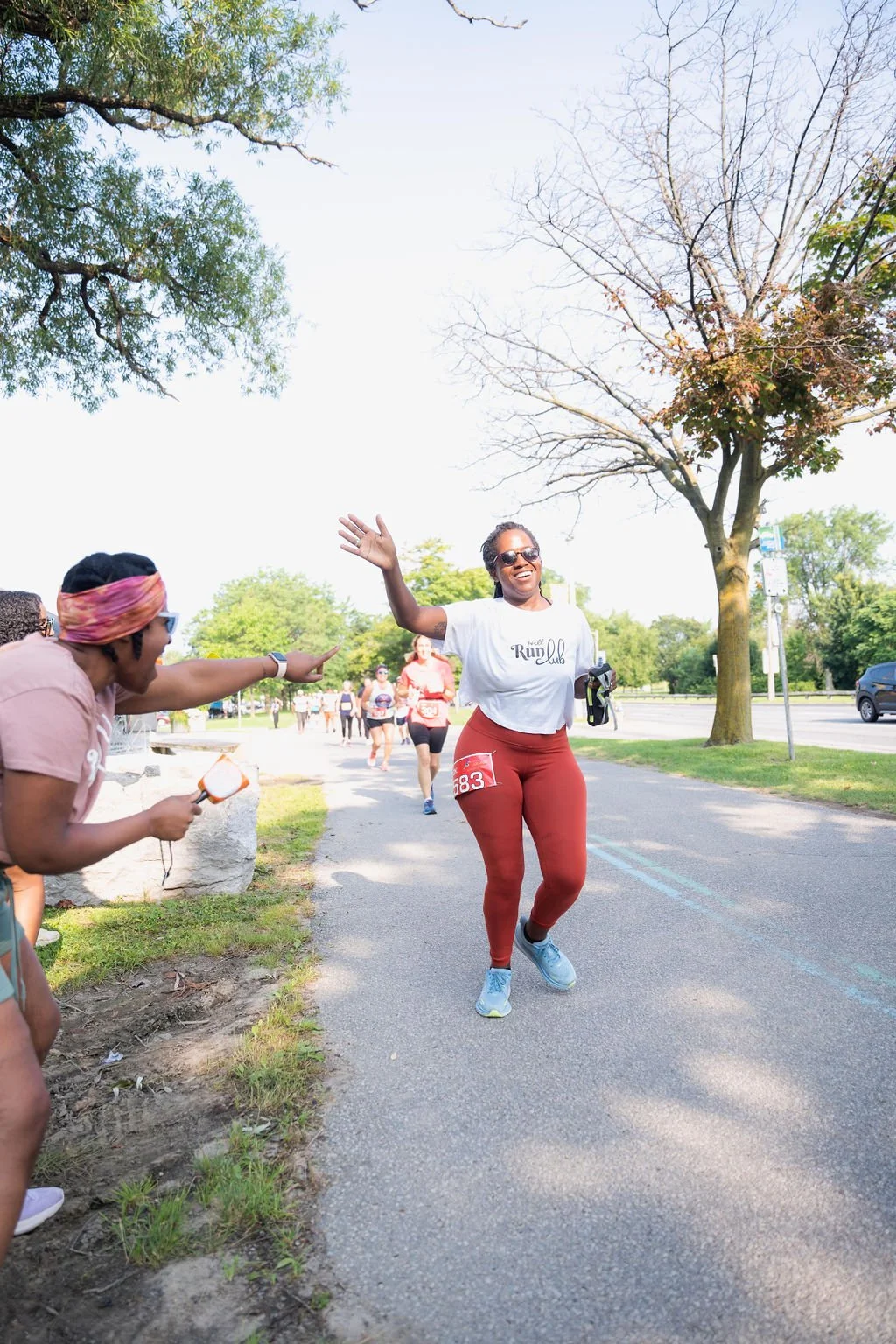 A woman in athletic wear and sunglasses celebrates while running in a race, with a smile and one arm raised, as volunteers cheer her on at a marathon event.