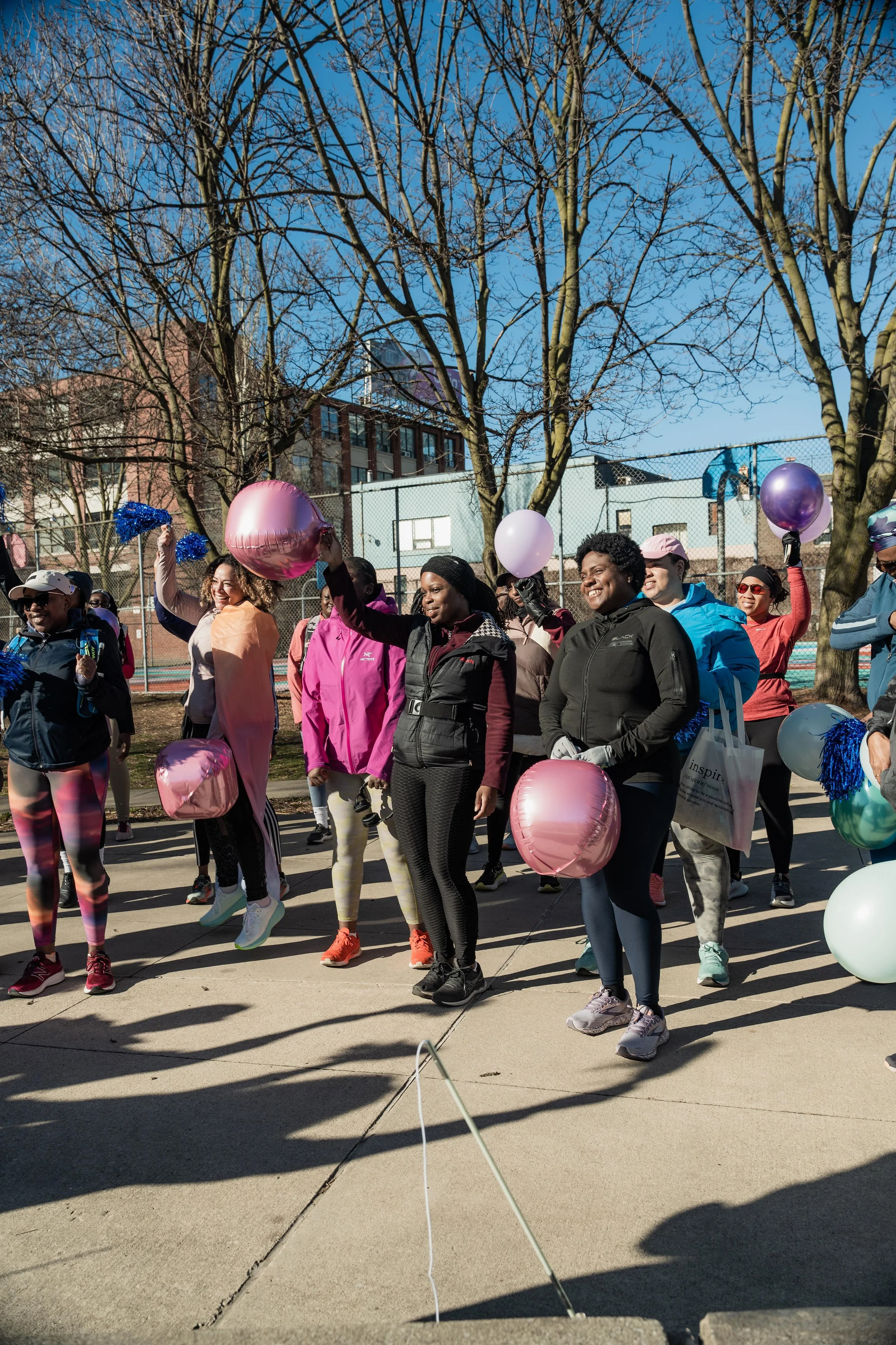 A group of diverse women participating in an outdoor event, holding colorful balloons and cheering, on a sunny day with leafless trees and a sports court in the background.