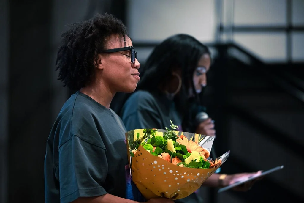 A woman with glasses holding a bouquet of colorful flowers during a ceremony or event, with another woman speaking into a microphone in the background.