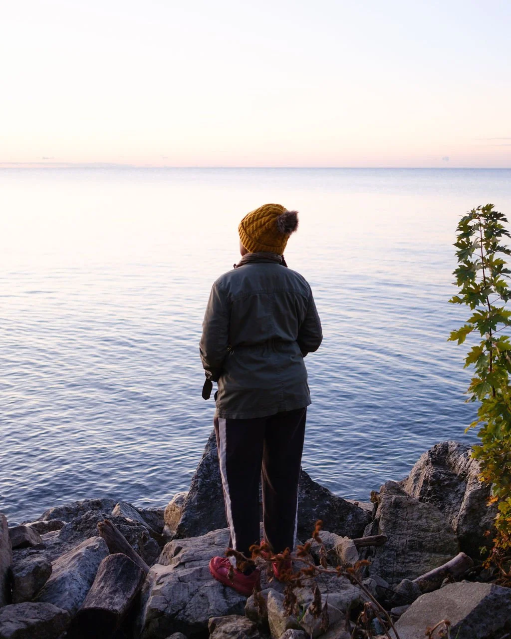 Person standing on rocks by the water, facing the ocean at sunset or sunrise, wearing a mustard beanie, dark jacket, and red shoes.