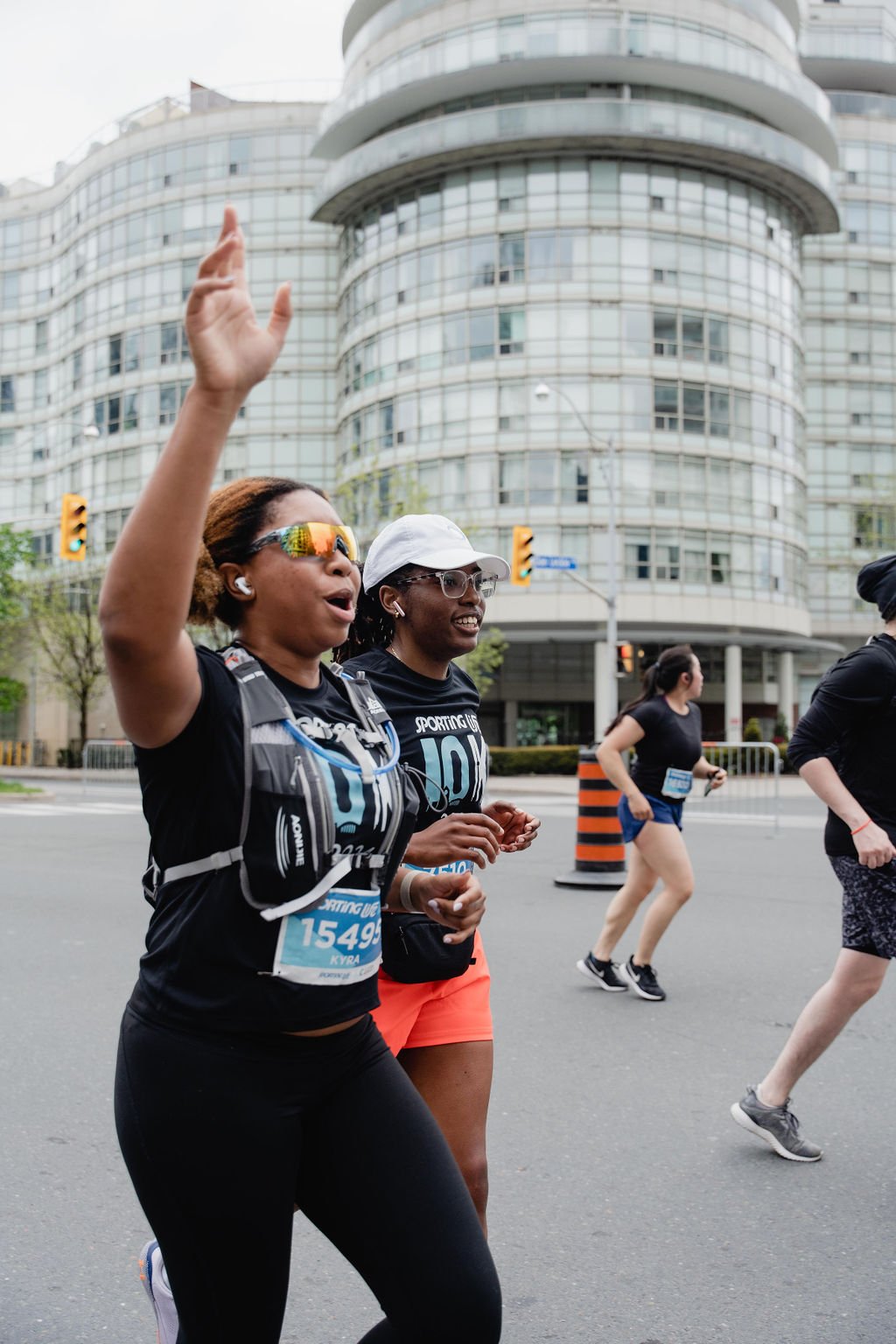 Two women running in a street race, one is waving with her hand, both smiling, with other runners nearby, tall modern building in the background.