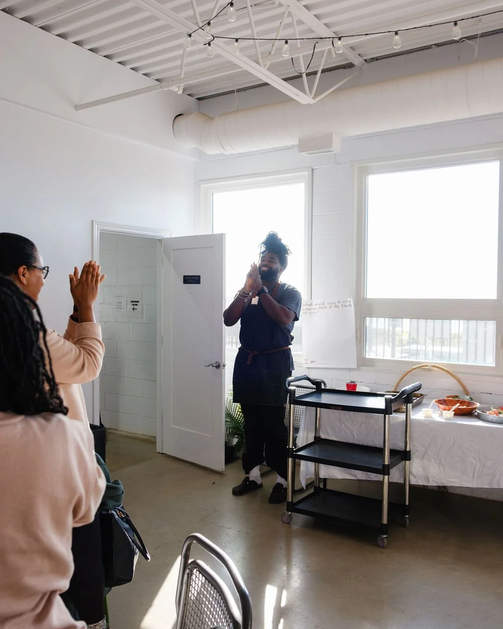 A man with dreadlocks and beard smiling with hands clasped in front of two women in a bright room. One woman is wearing glasses and tan sweater, clapping. The room has large windows, white walls, and a table with snacks.