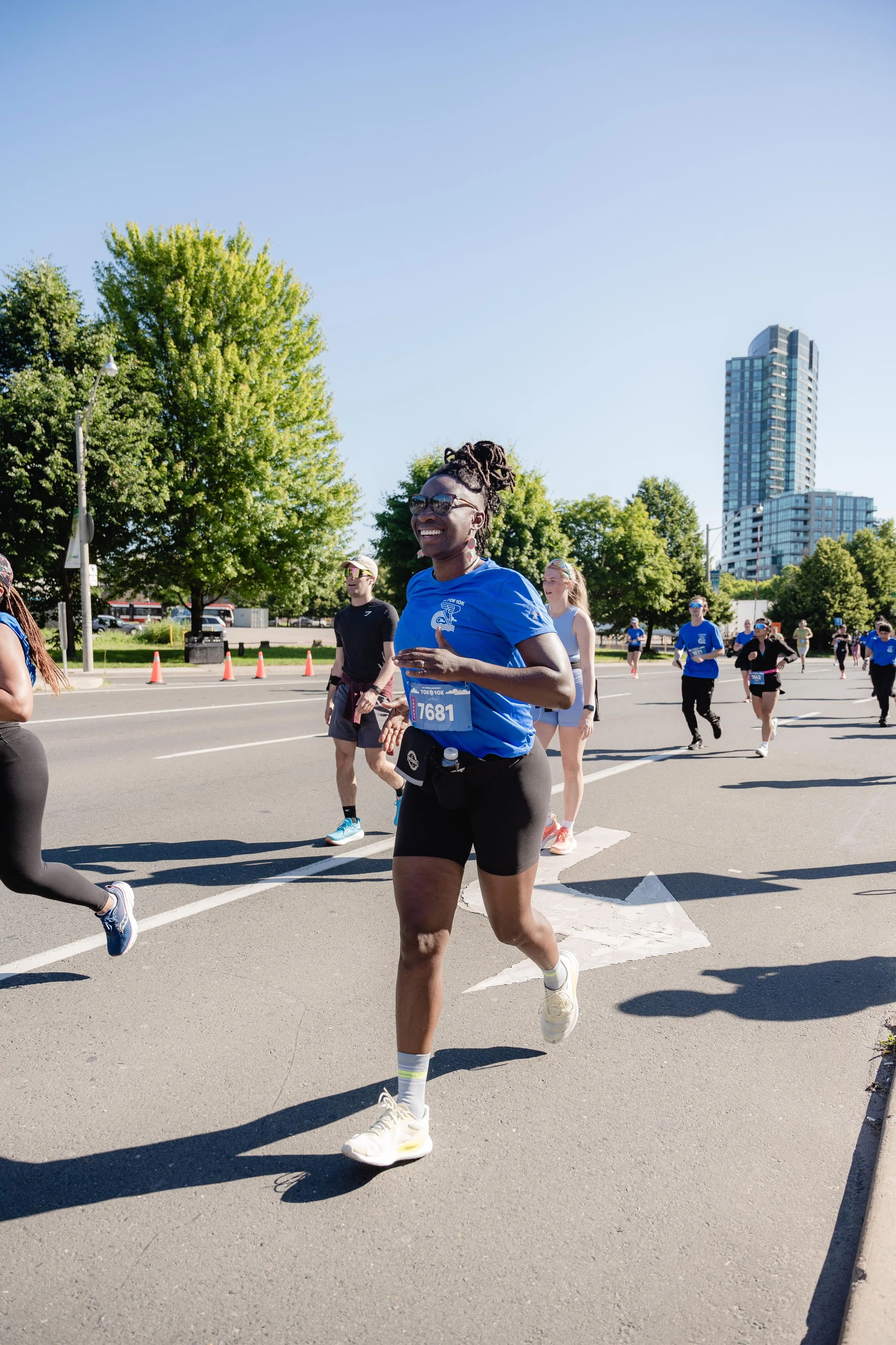 A group of people participating in a marathon or running event outdoors on a sunny day, with a tall building and green trees in the background. The woman in front is smiling, wearing sunglasses, a blue shirt, black shorts, and white running shoes.