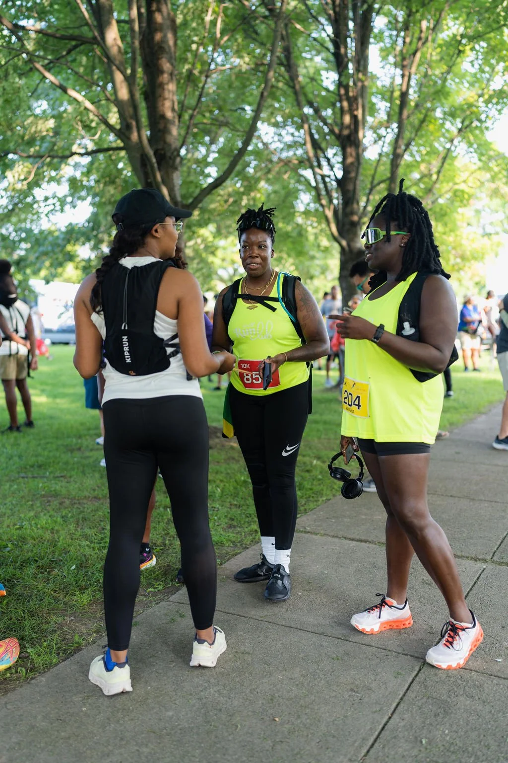 Three women in athletic gear having a conversation at an outdoor event, with trees and other participants in the background.