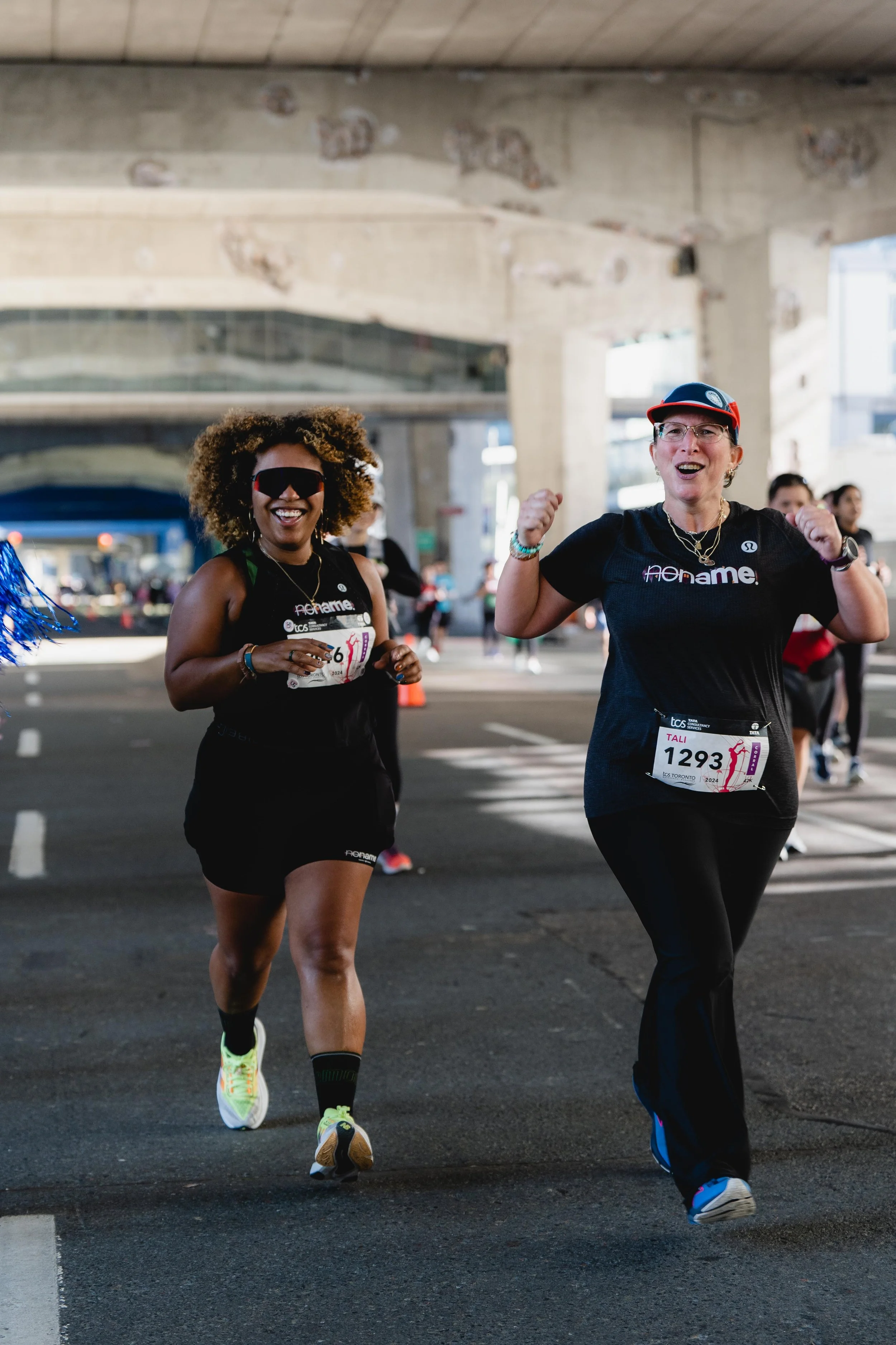 Women running in a marathon, smiling and celebrating under a bridge.