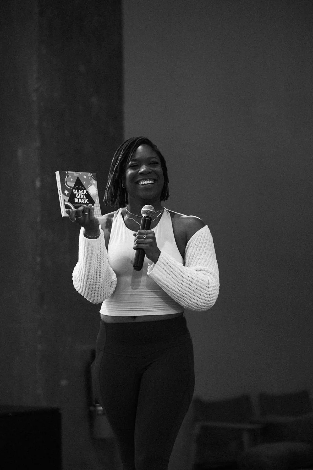 A woman with curly hair smiling while holding a book titled 'Black Girl Magic' in her left hand and a microphone in her right hand, standing in a dimly lit room with chairs in the background.