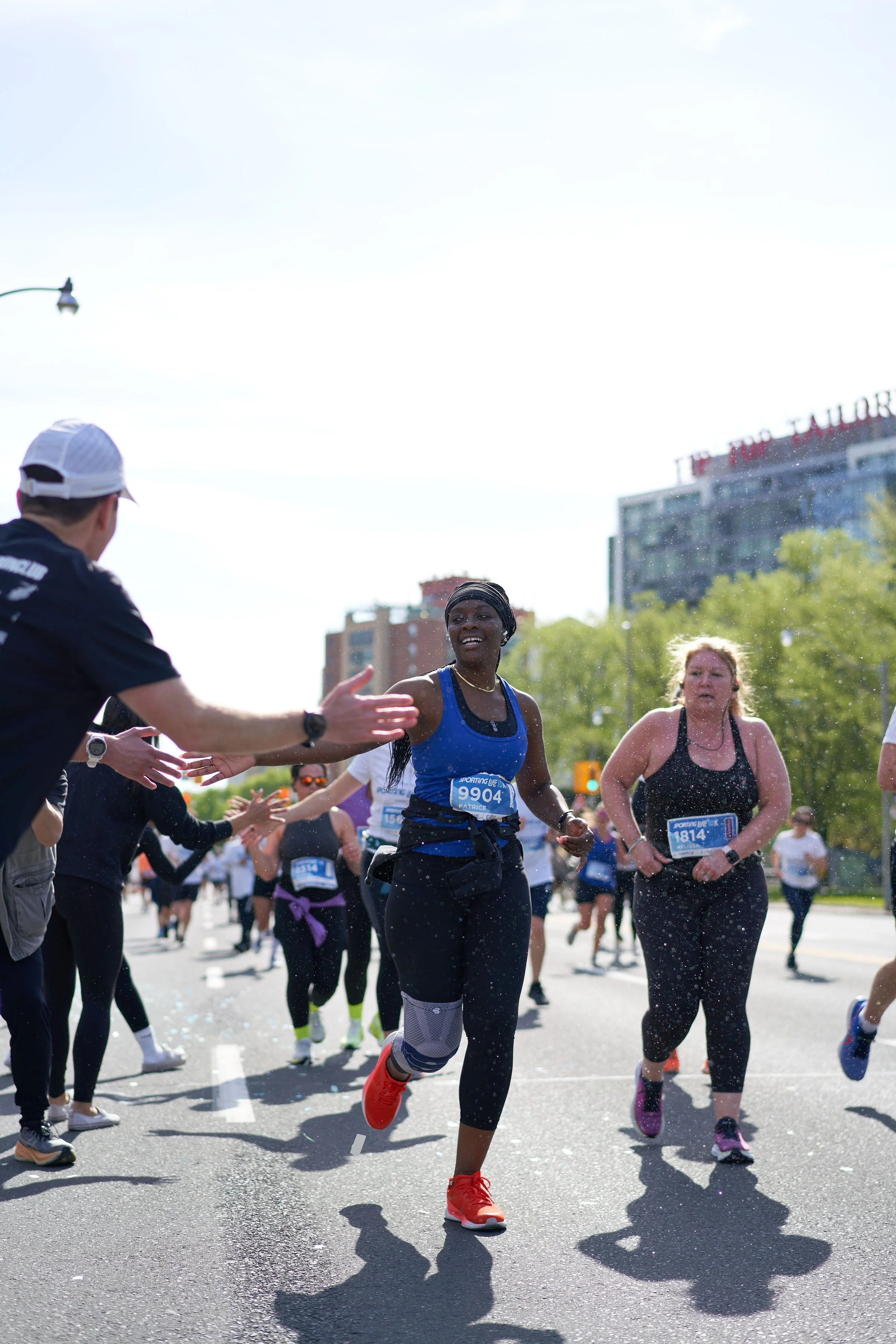 Runners for Hill Run Club participating in a 10K race on a city street, with a woman in a blue tank top and black leggings giving high-fives to spectators.