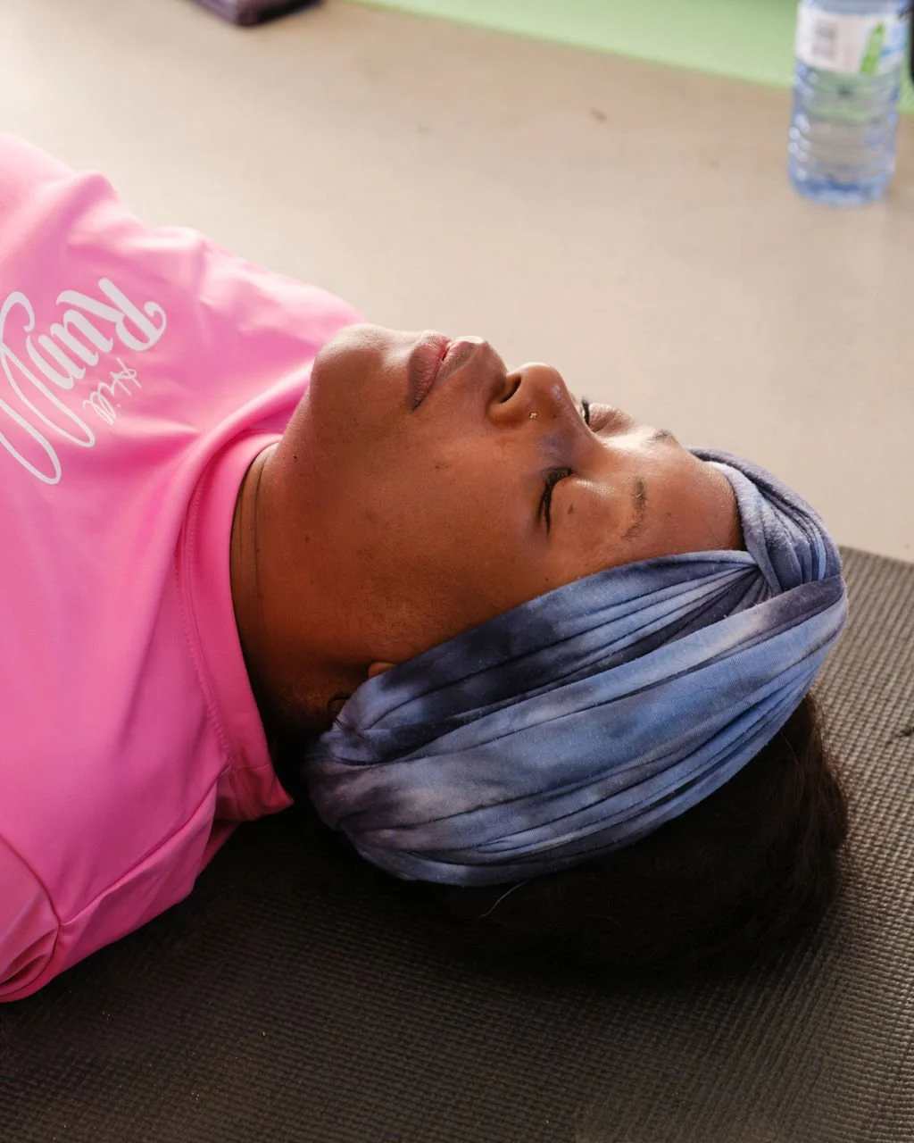A woman with a blue tie-dye headwrap and a pink shirt lying down on a brown mat with her eyes closed.