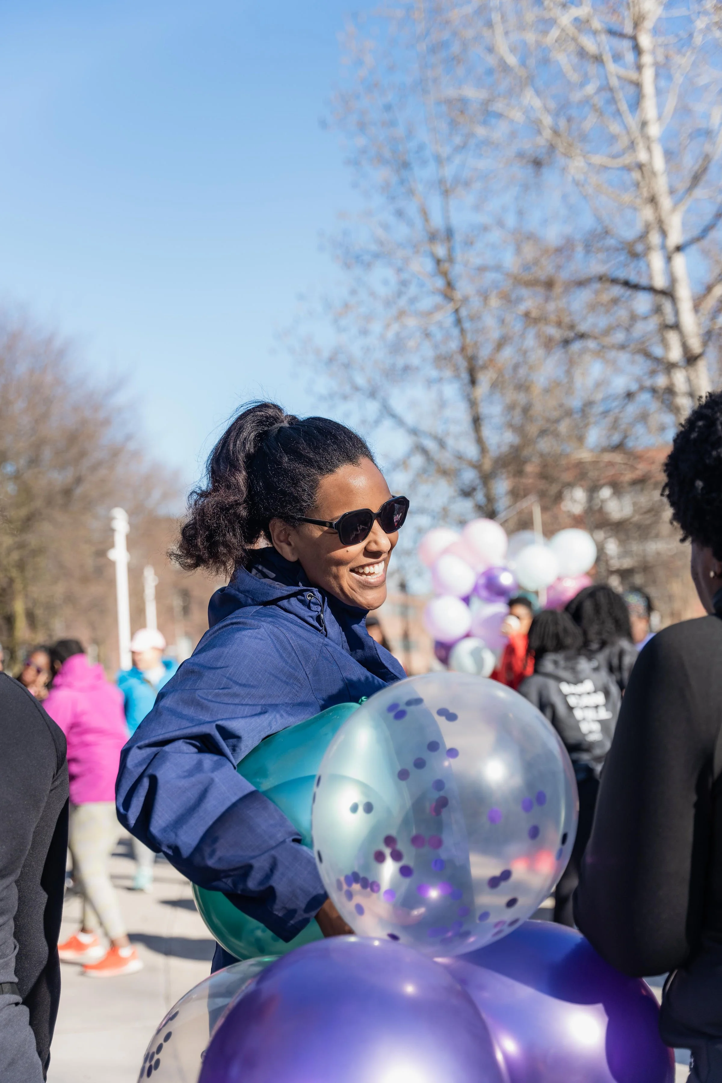 A woman with sunglasses and a blue jacket holding teal and clear balloons with purple confetti at an outdoor event on a sunny day.