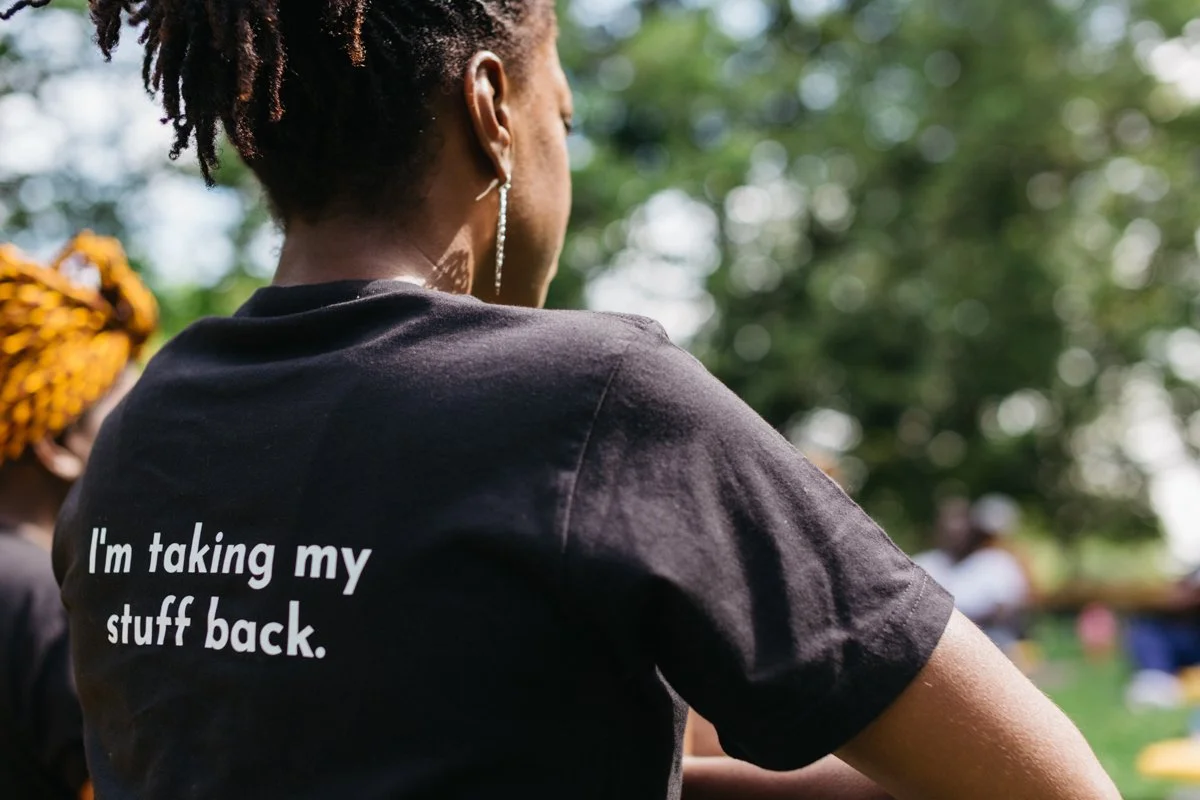 Back of woman wearing black T-shirt with white text that says, "I'm taking my stuff back," outdoors with trees and other people in the background.