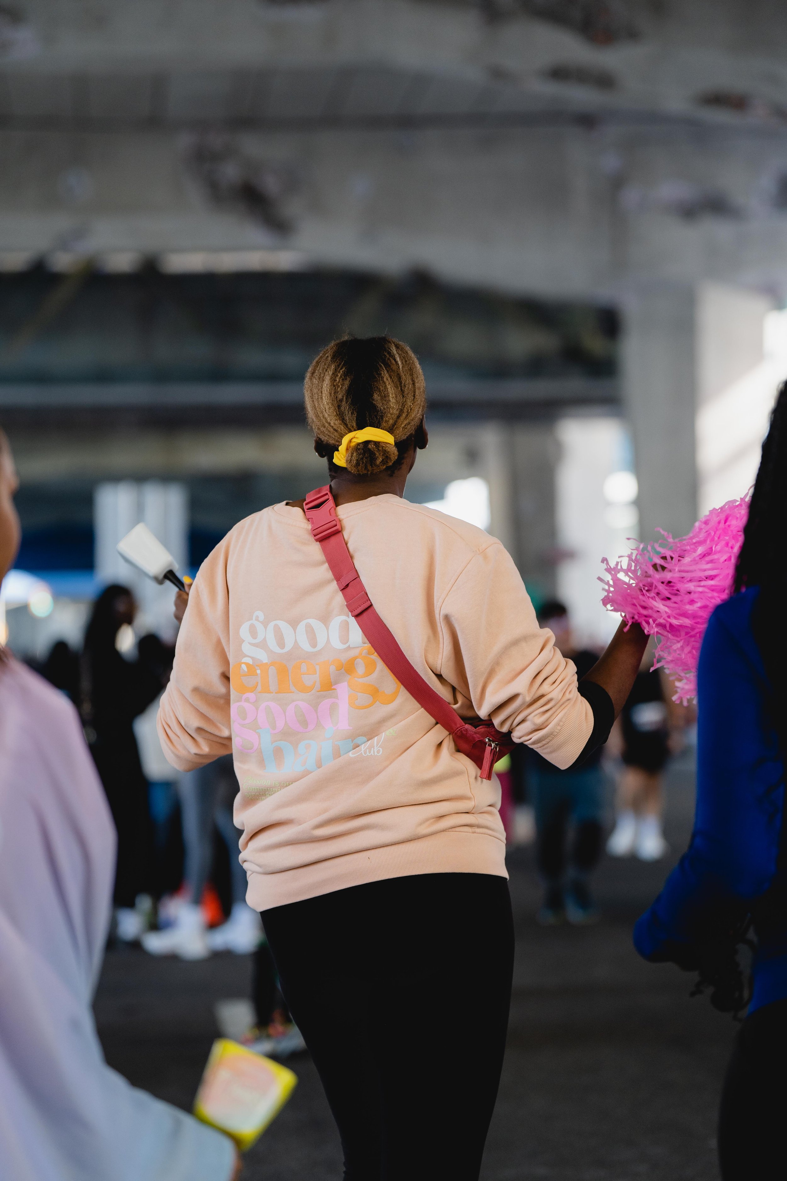 A woman with a yellow hair tie and beige sweatshirt walking under an overpass with a crowd in the background.