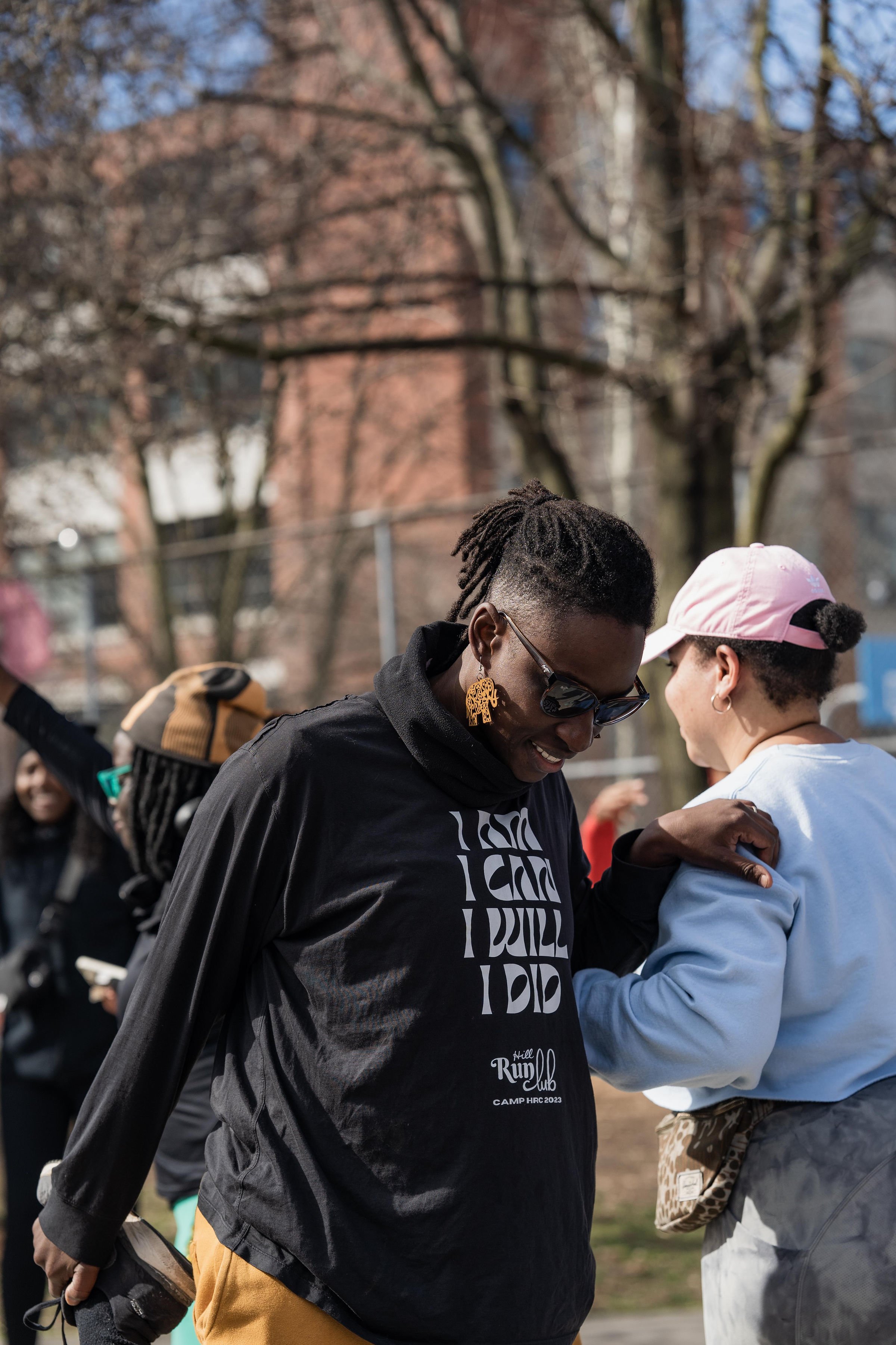 Two women sharing a joyful moment outdoors, one wearing sunglasses and a black jacket, the other wearing a pink cap and white sweater, with trees and buildings in the background.