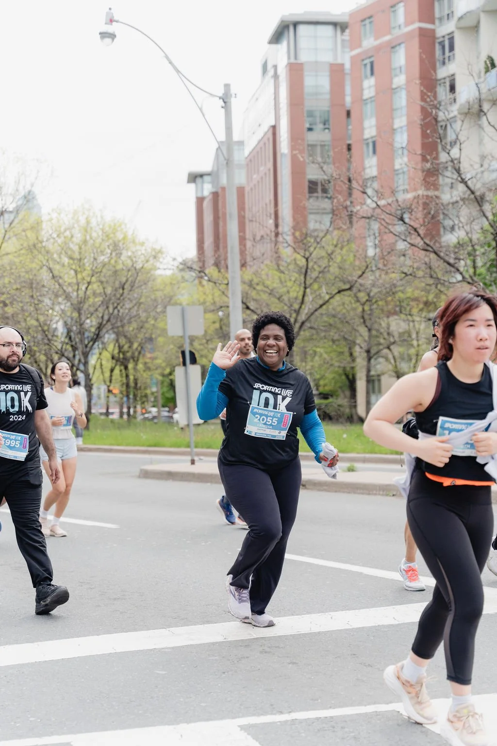 Group of people participating in a running event on a city street, smiling and waving, with trees and high-rise buildings in the background.