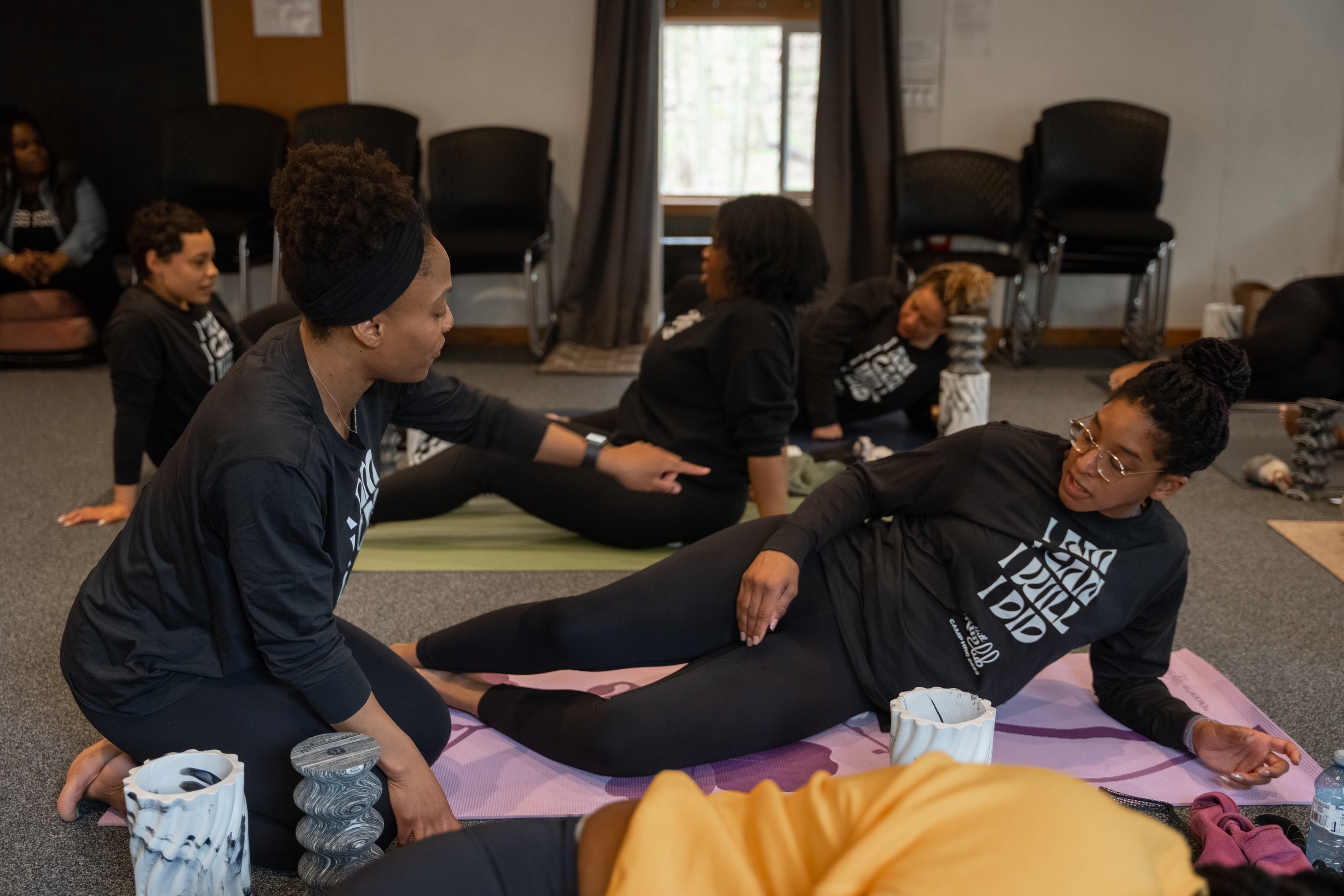 Women in black shirts practicing yoga on mats, one guiding another during a massage or stretching session inside a room.