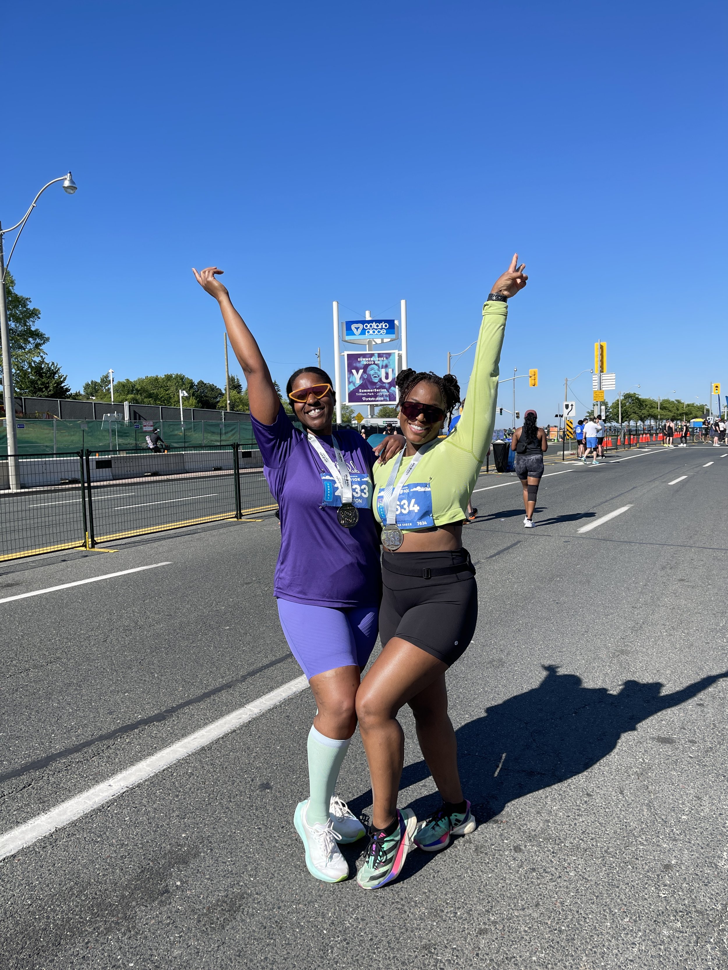 Two women celebrating after a race, wearing medals and race bibs, outdoors on a sunny day with a blue sky.