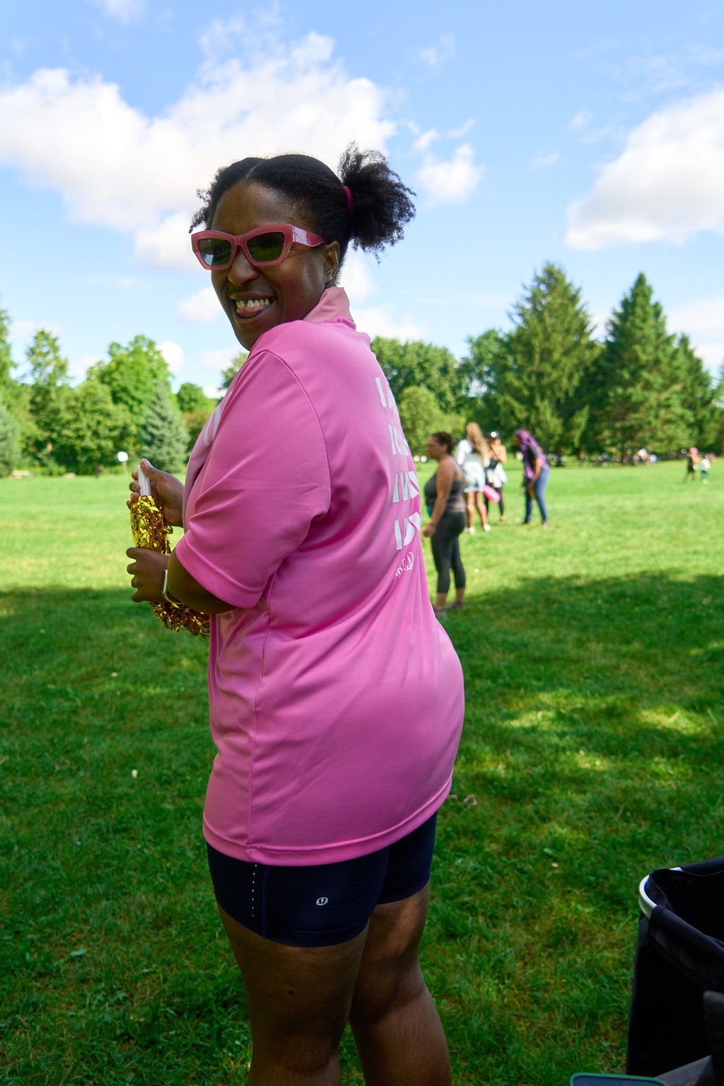 A woman in a pink jacket and black athletic shorts standing outdoors on a grassy field with trees and blue sky, smiling and holding some gold-colored objects, with a group of people in the background.