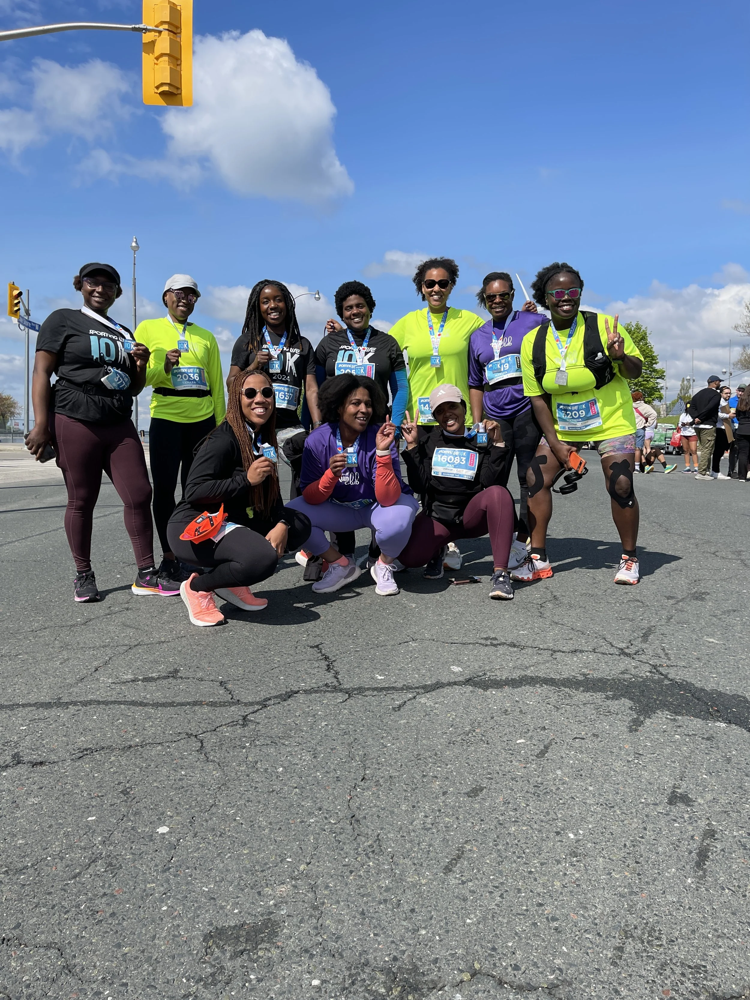Group of eleven diverse women participants in a race or marathon, wearing athletic clothing with race bibs and medals around their necks, posing outdoors on a cloudy day.