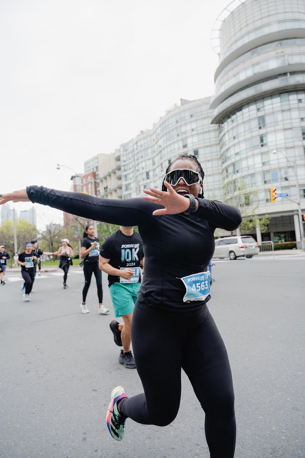 A woman participating in a marathon, wearing sunglasses and black athletic clothing, is running with her arms outstretched in an urban area with modern buildings in the background.