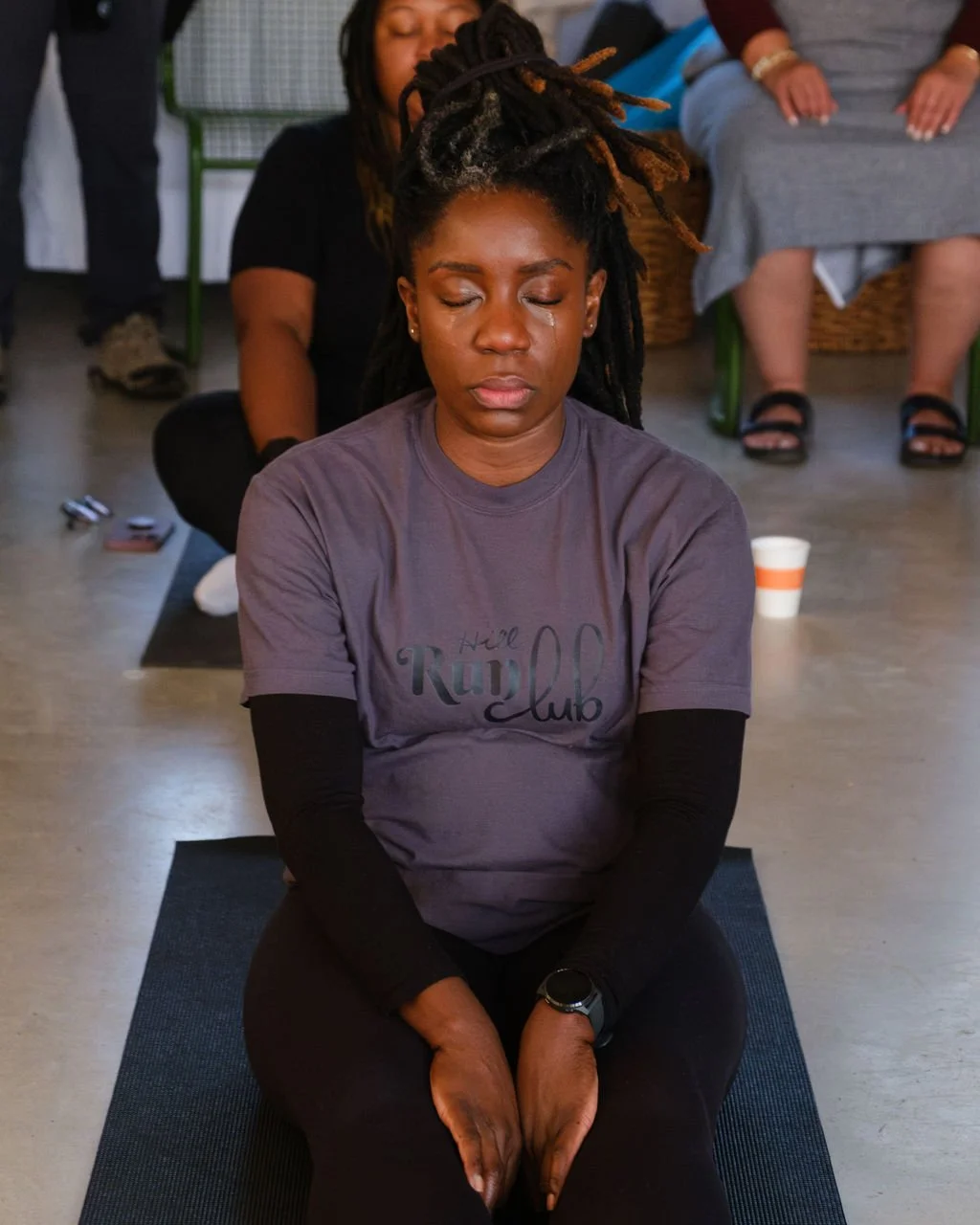 A woman with dreadlocks and tears on her face, practicing yoga or meditation in a group, sitting cross-legged with hands resting on her feet, in an indoor setting.