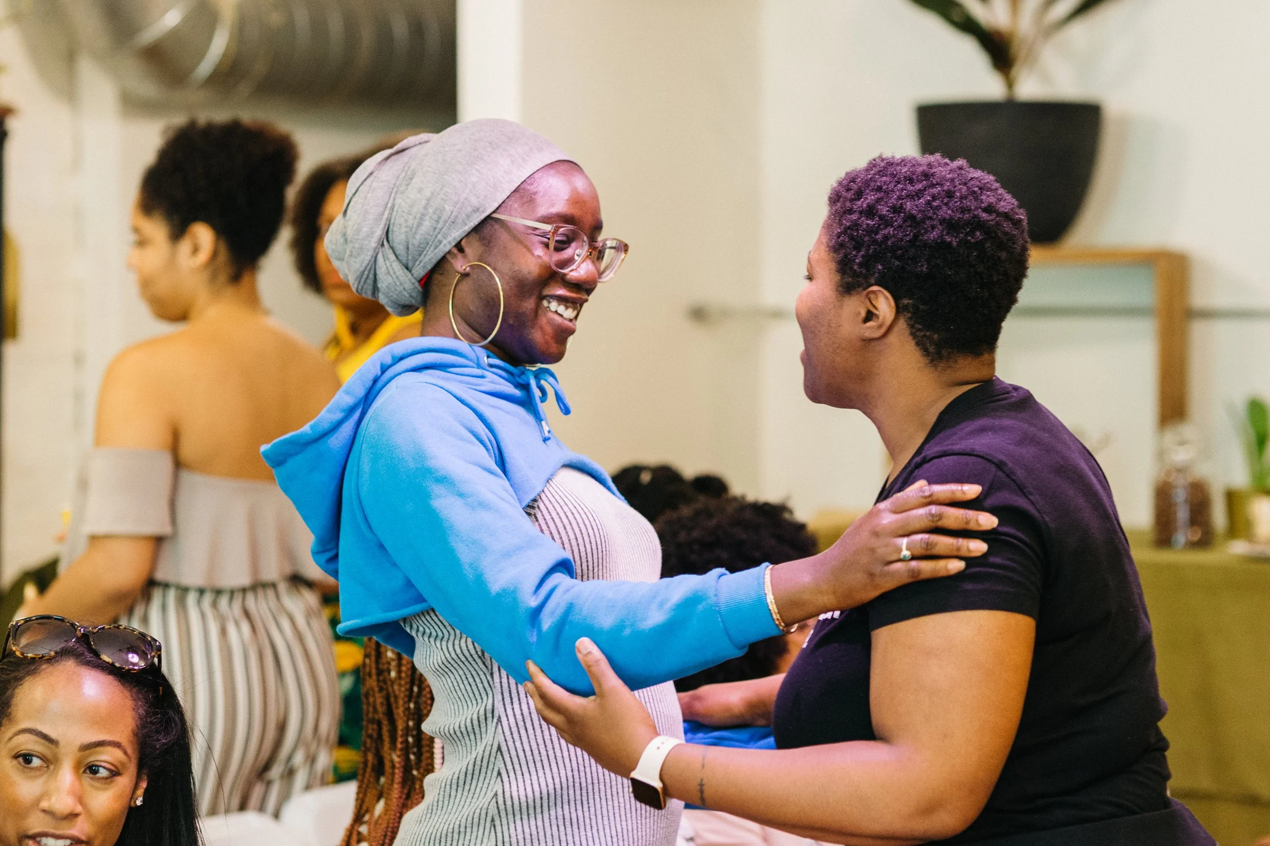 Two women are smiling and holding each other at a gathering. One woman wears glasses, a grey headscarf, a blue hoodie, and striped dress, while the other woman has short purple hair and a black shirt. There are other people and decorative items in th