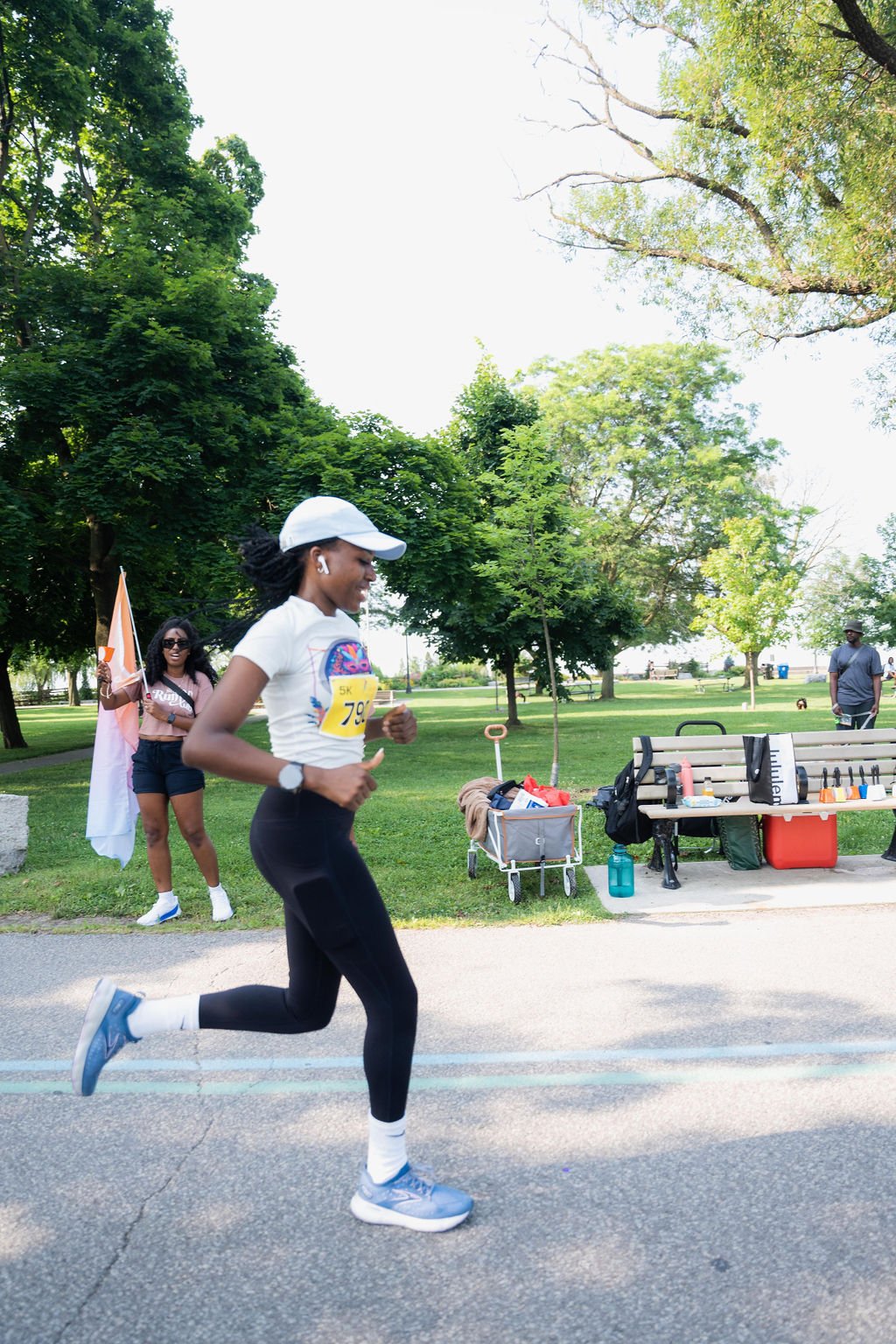 A woman running in a park during a race, wearing a white cap, white t-shirt, black leggings, and blue athletic shoes. She has a race bib on her shirt labeled '5K 79'. In the background, another woman is holding a flag and smiling, and a man is standi