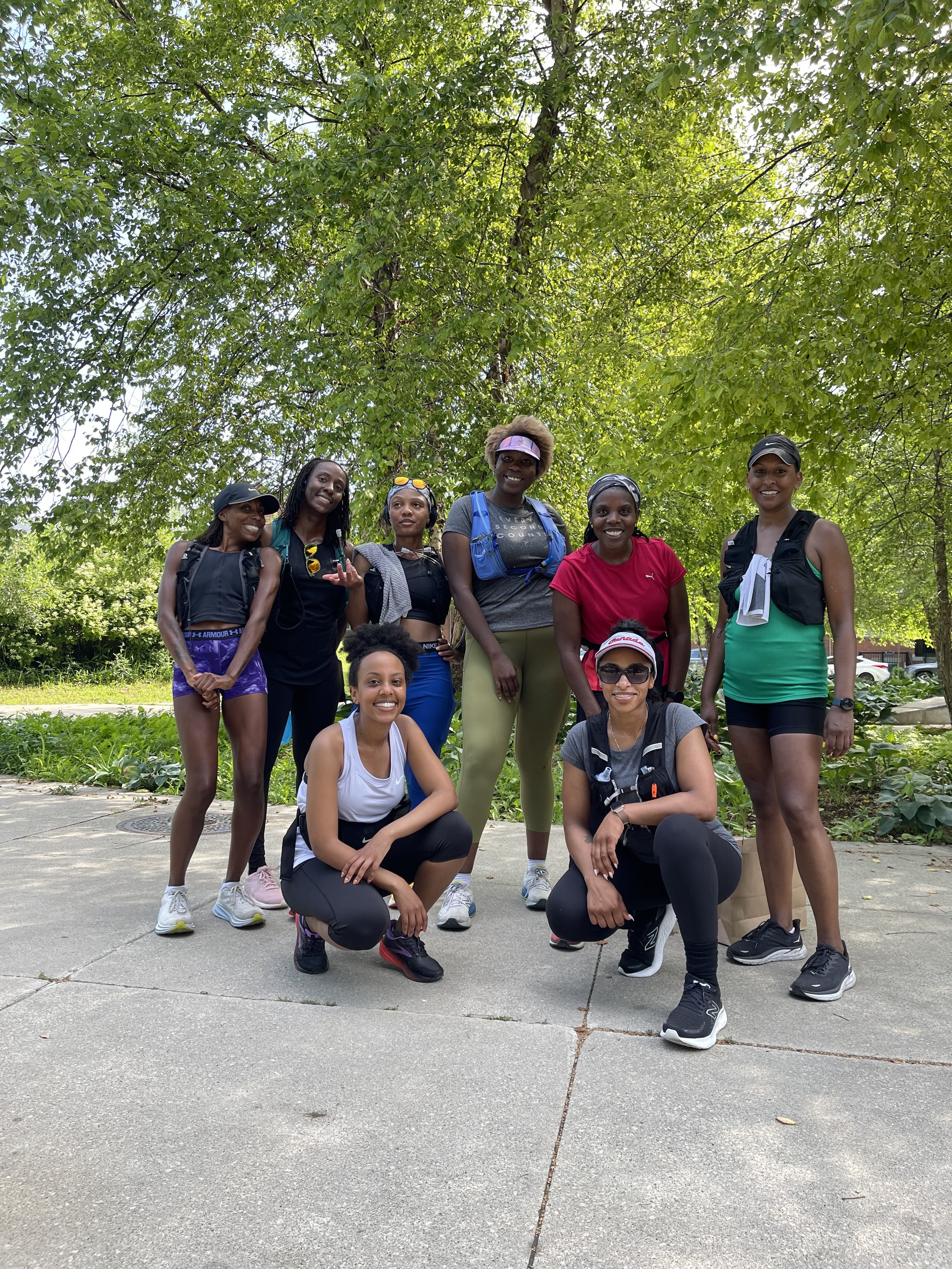 Group of eight women in athletic wear and backpacks outdoors on a sidewalk under a large leafy tree, smiling and posing for a photo.