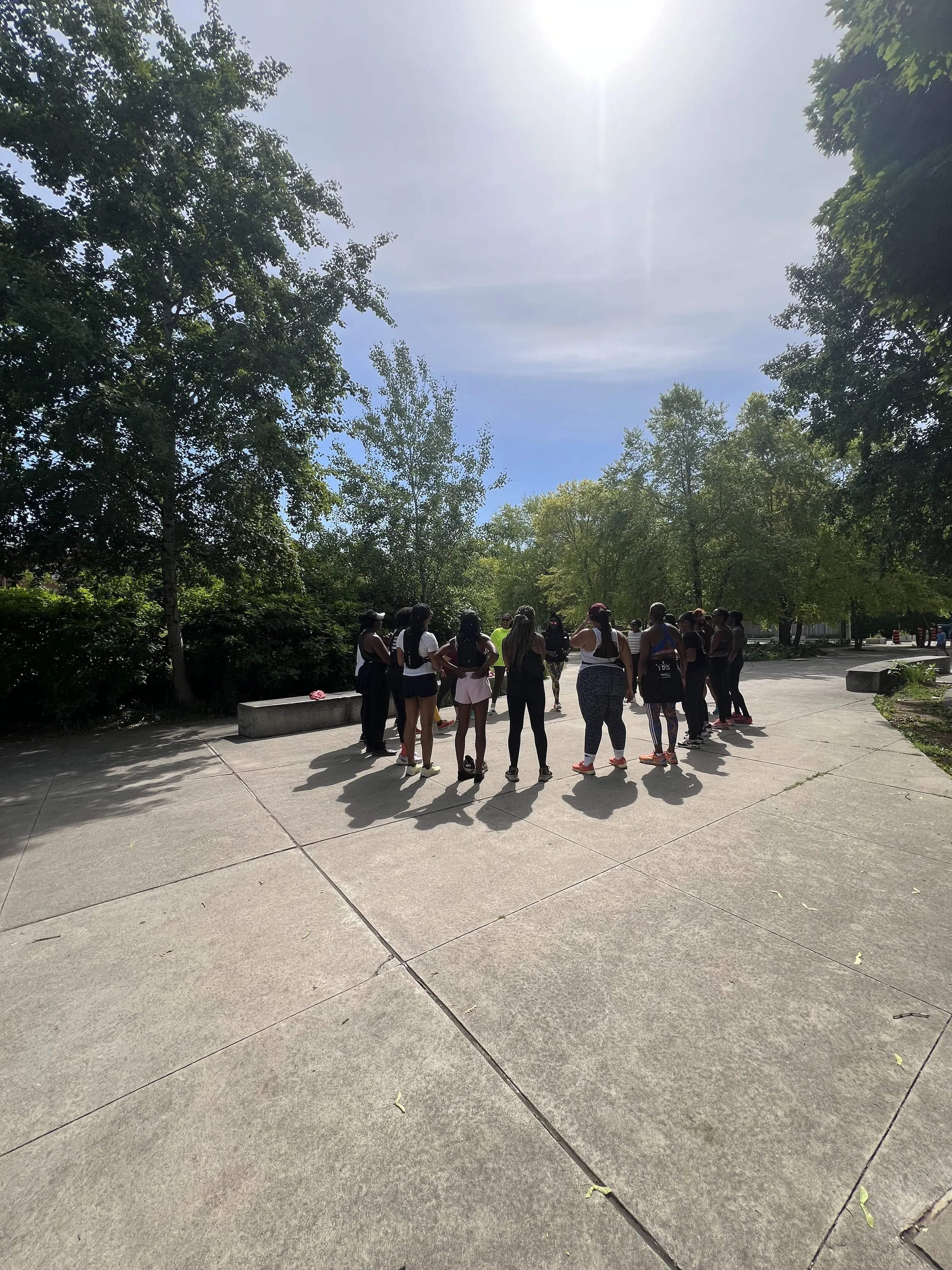A group of people standing in a circle outdoors on a paved area surrounded by trees on a sunny day.