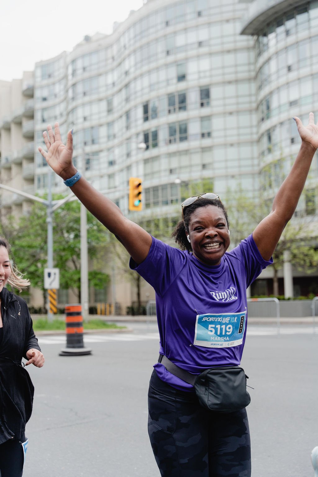 Happy woman running in a race with arms raised, wearing a purple shirt, race bib with number 5119, and sunglasses on head, in an urban setting.