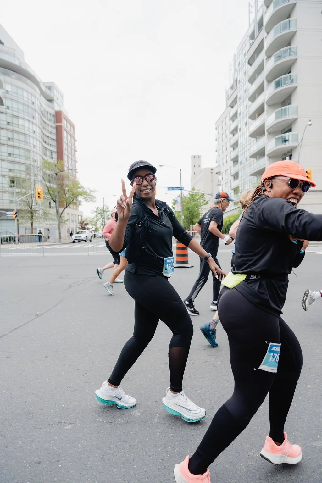 Smiling woman in black athletic attire posing with a peace sign during a road race in an urban area, with other runners and modern buildings in the background.