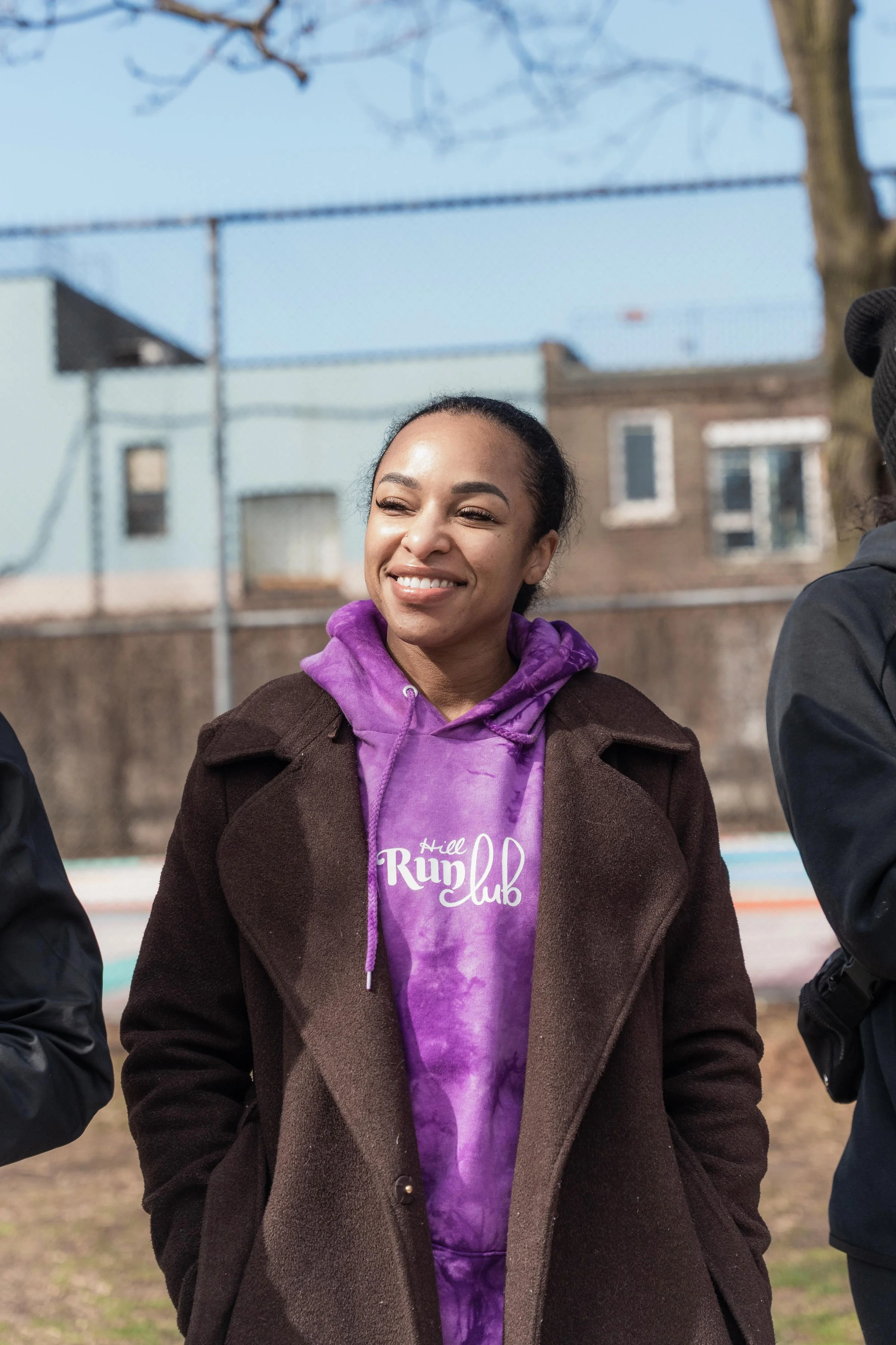 A smiling woman wearing a purple tie-dye hoodie with 'Hill Run Club' written on it, standing outdoors on a sunny day with a chain-link fence and buildings in the background.