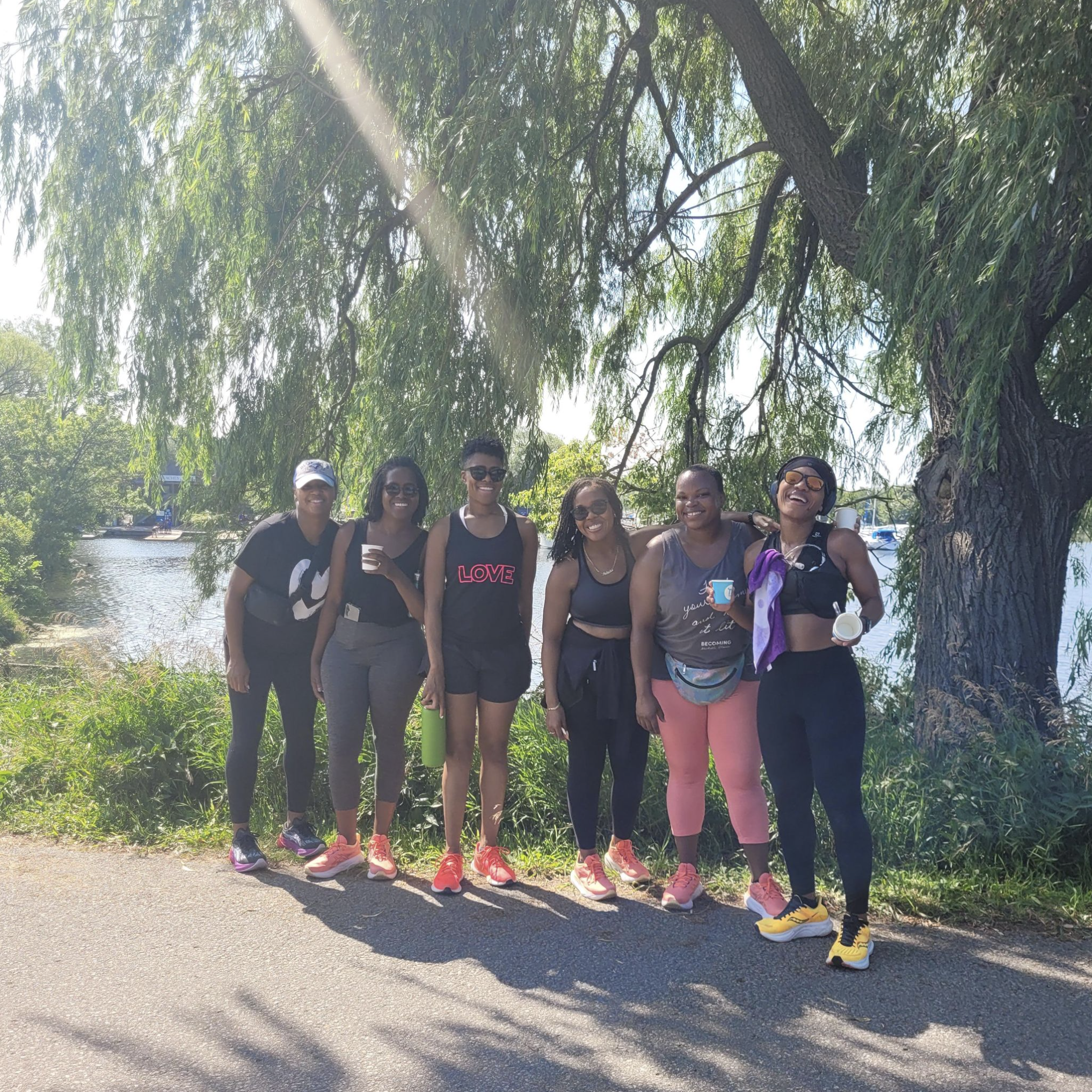 Six women stand together outdoors beside a river, with trees and greenery in the background, smiling and posing for a photo on a sunny day.