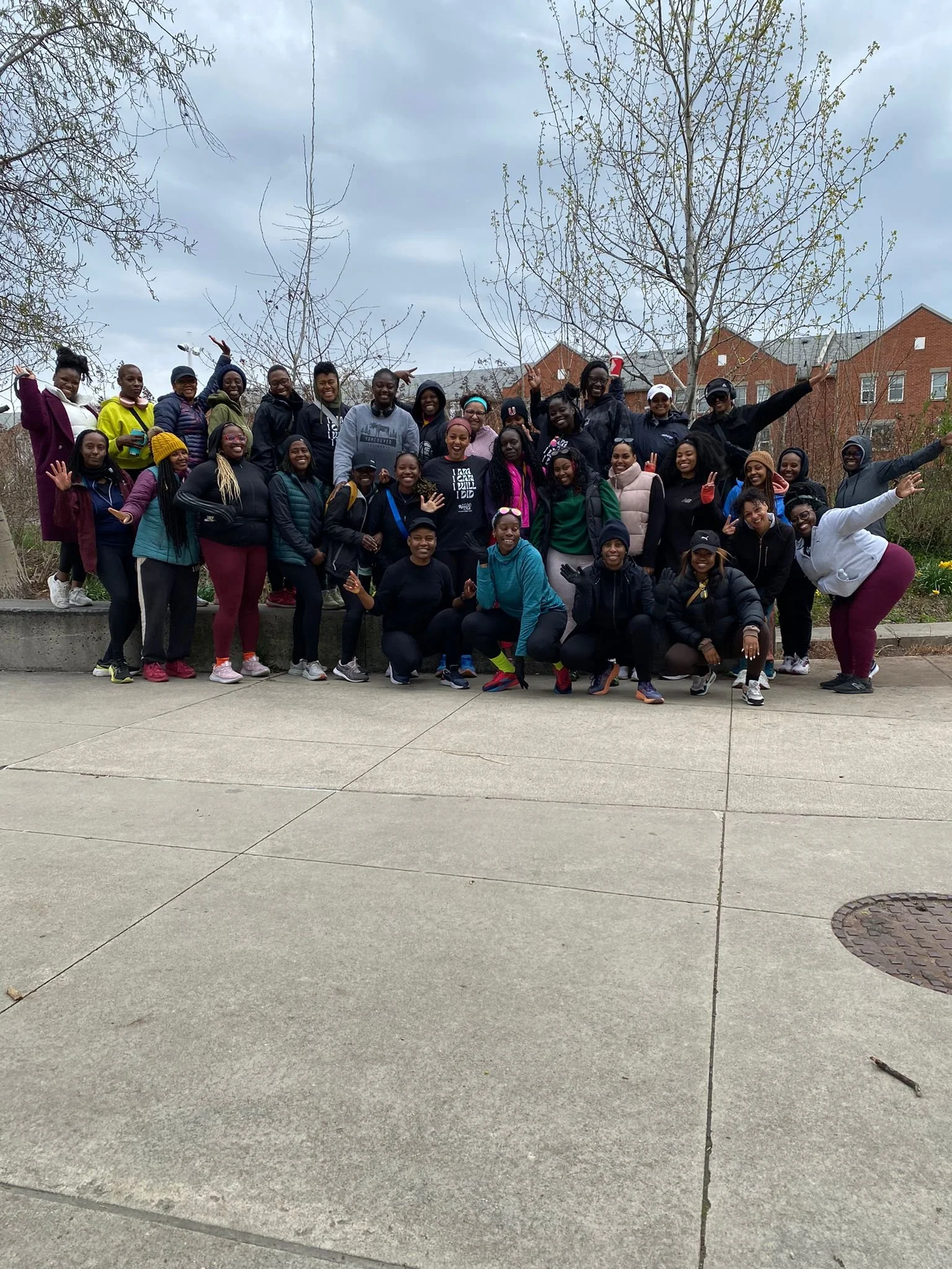 Group of smiling women and girls posing outdoors on a sidewalk with leafless trees and brick buildings in the background.