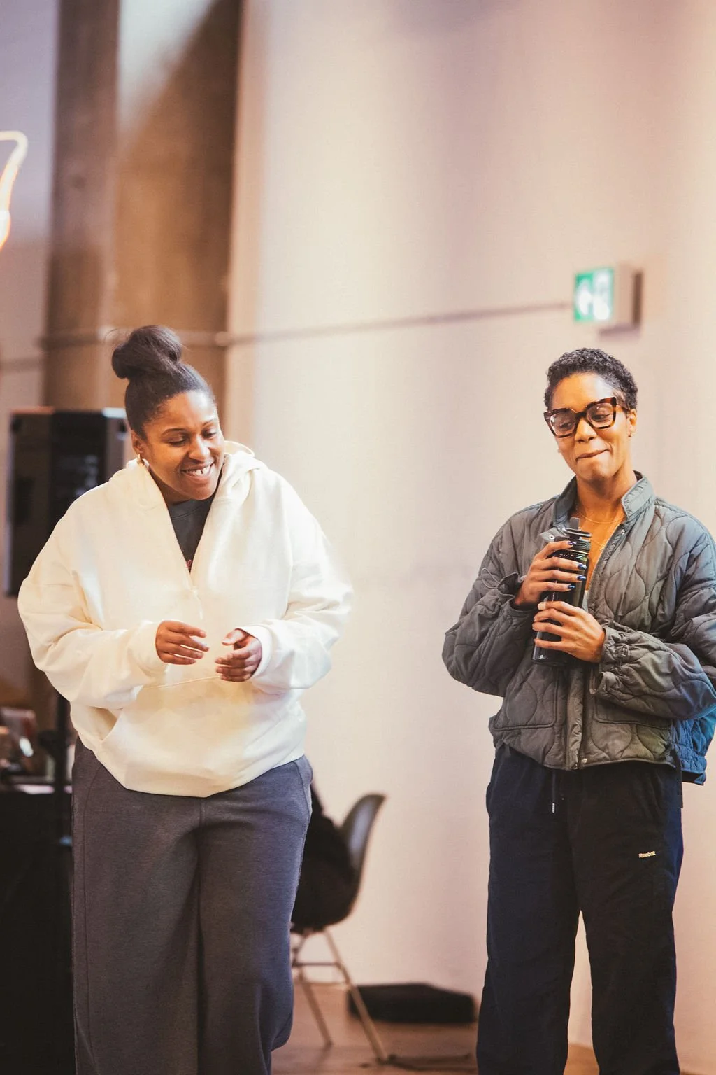 Two women are standing and smiling, one is wearing a white hoodie and the other is wearing a dark jacket and glasses, inside a room with beige walls.