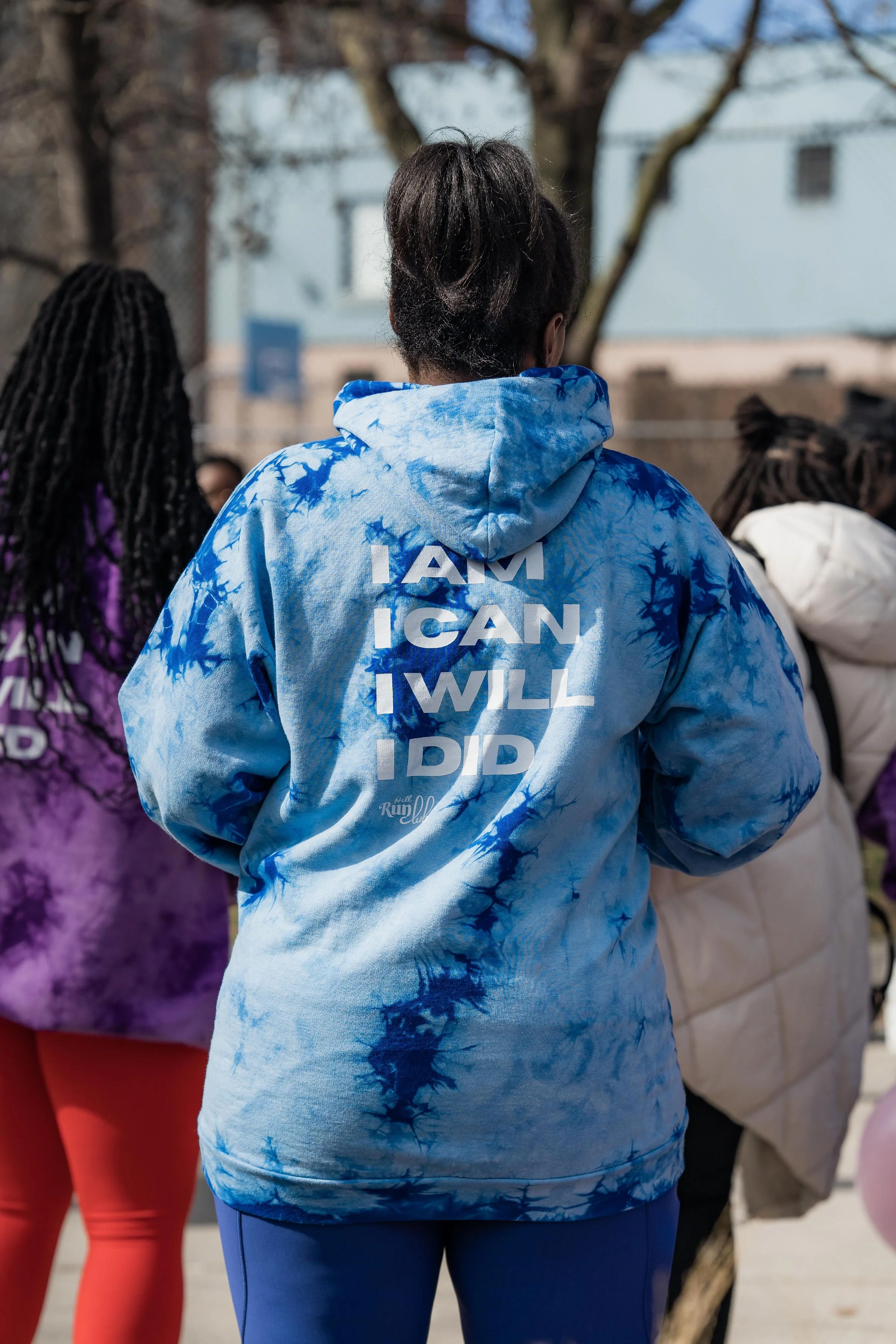 Back of a person wearing a blue tie-dye hoodie with the words 'I AM I CAN I WILL I DID' printed on it, standing among a group of people outdoors.